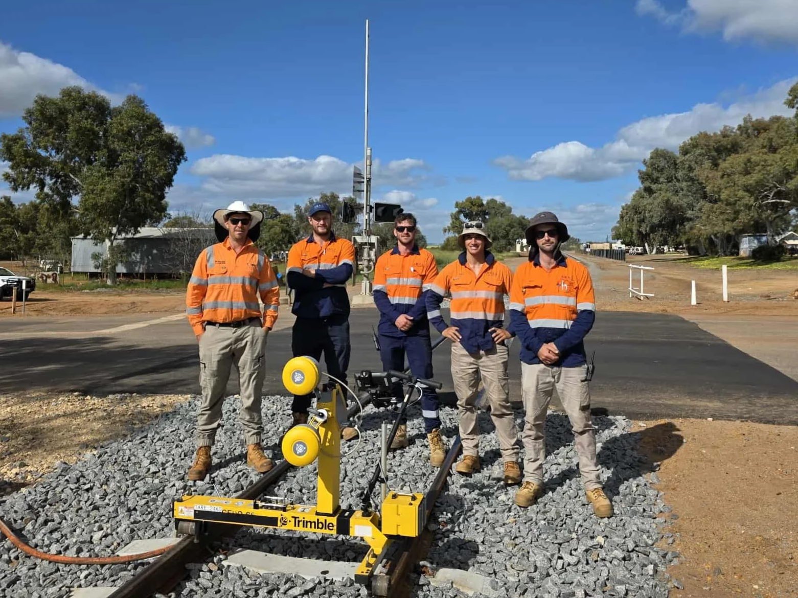 Five construction workers in orange safety vests stand on a gravel surface near track switch equipment outdoors on a sunny day with a blue sky and scattered clouds.