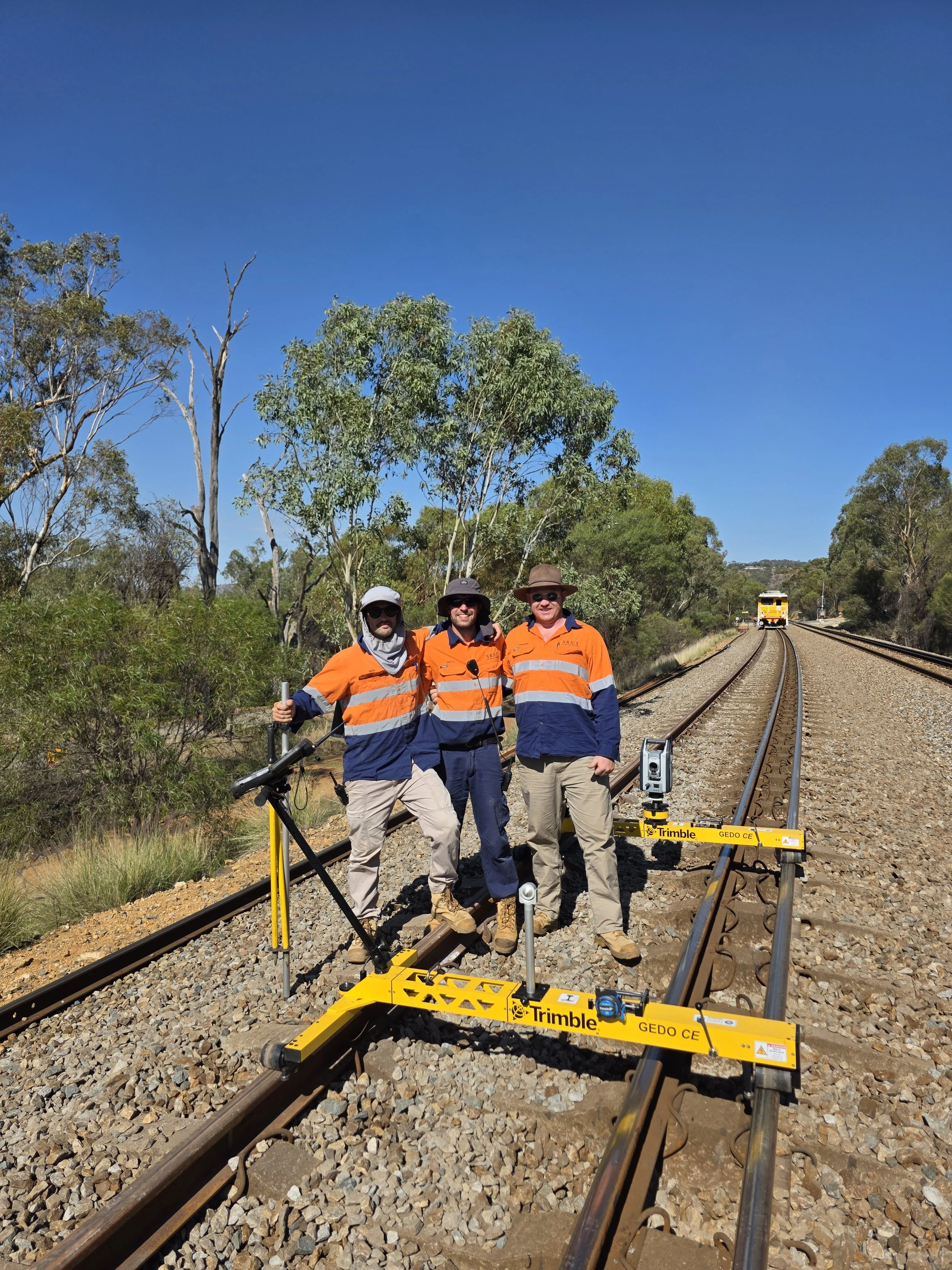 Three railway workers in orange and blue safety uniforms standing on train tracks with surveying equipment, surrounded by trees and a blue sky, with a train visible in the distance.