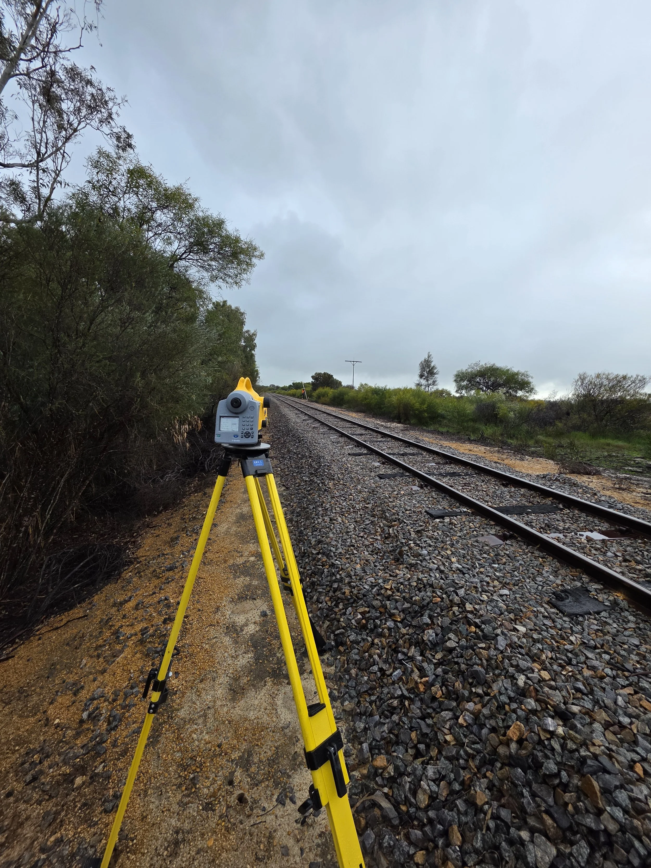 Surveying equipment on a tripod next to a railroad track on a cloudy day.