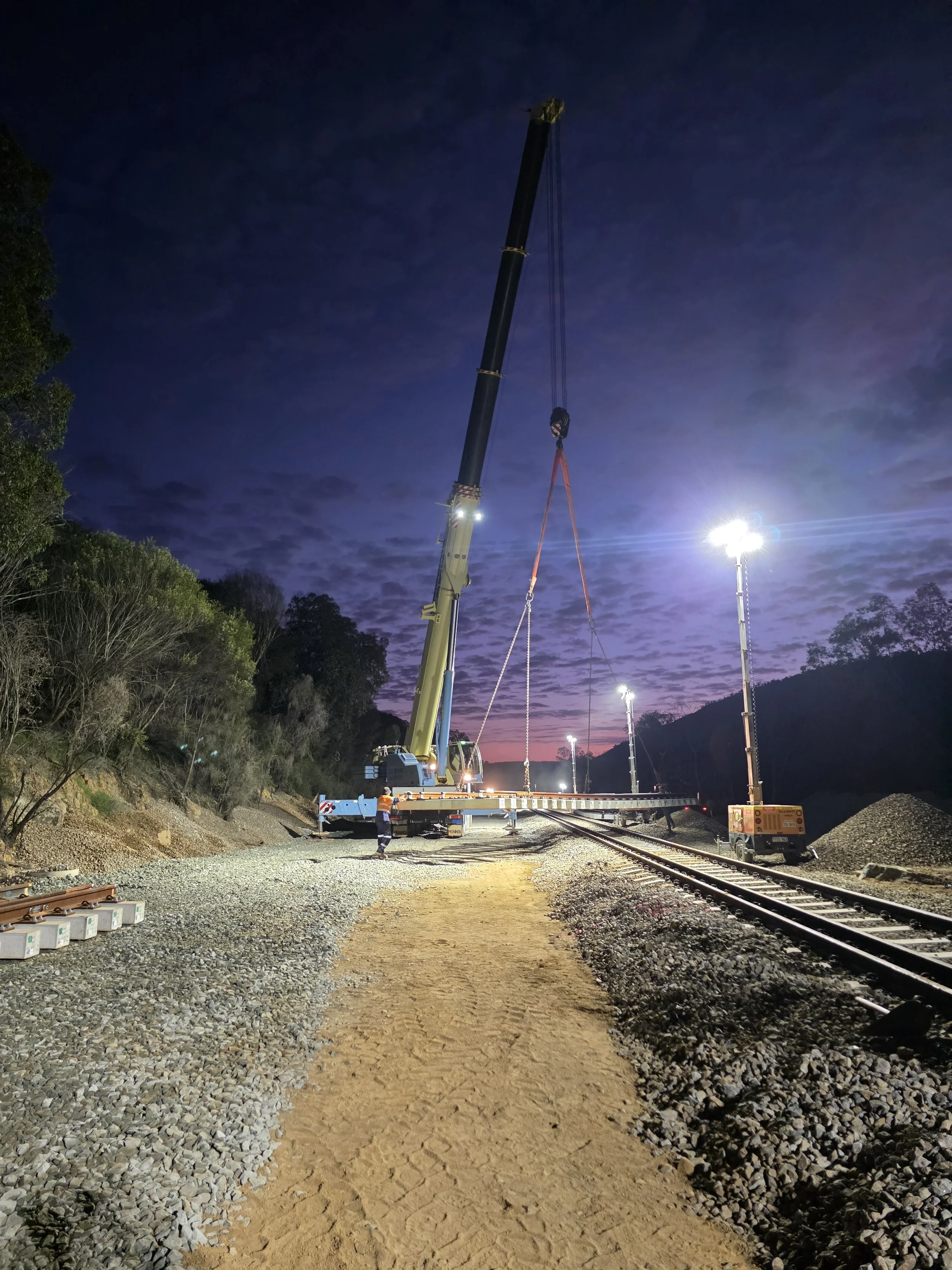 Workers operating a crane to install a new section of railway track at dusk, with mounted lights illuminating the construction site.