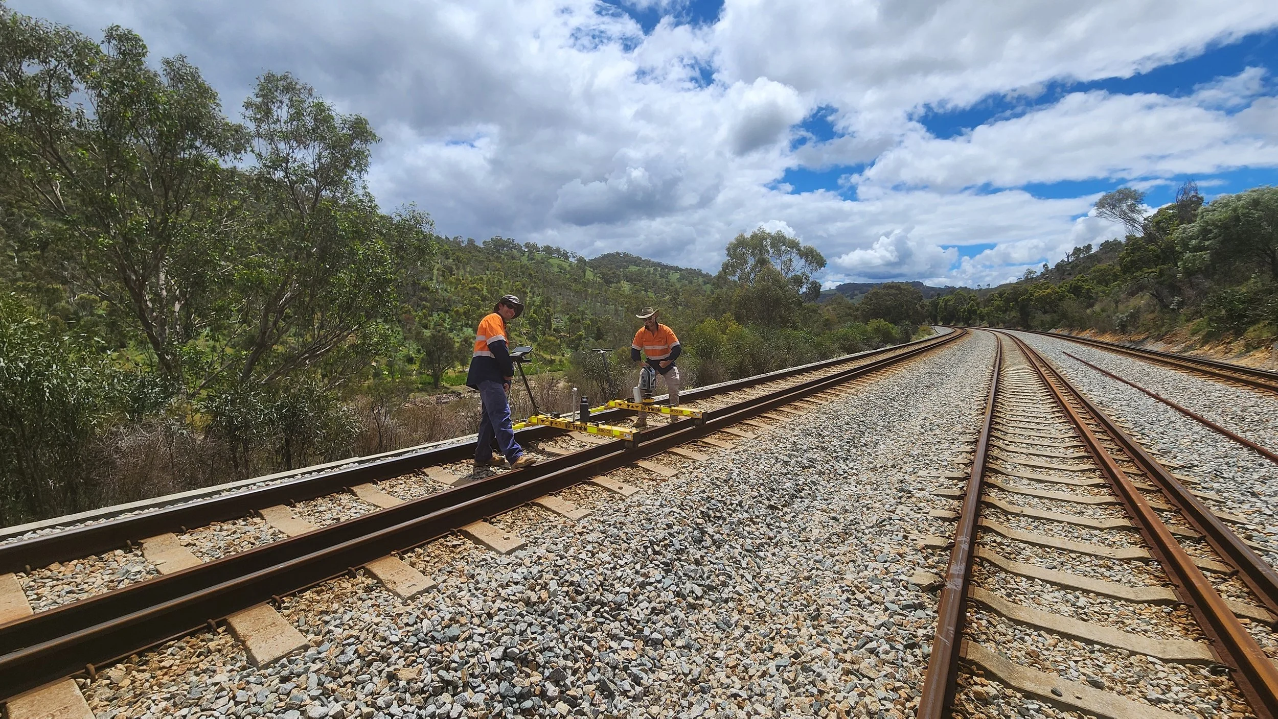 Two workers in safety vests working on a railway track in a rural area with trees and mountains in the background under a partly cloudy sky.