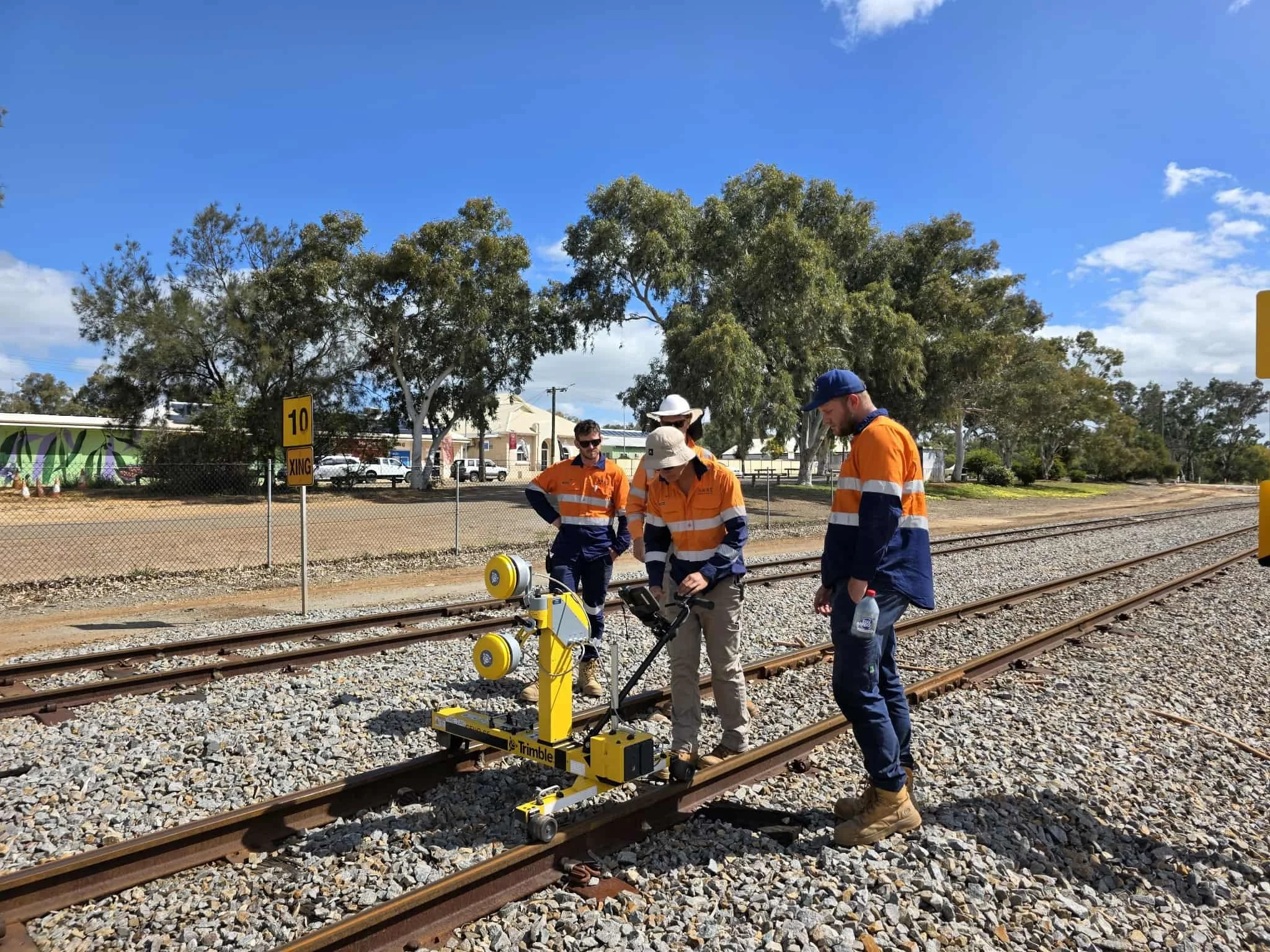 Four workers wearing orange and navy safety jackets, some with hats, are standing on railroad tracks inspecting equipment. Four large yellow devices are attached to a small yellow vehicle on the tracks. The background features trees, a building, and a clear blue sky with some clouds.