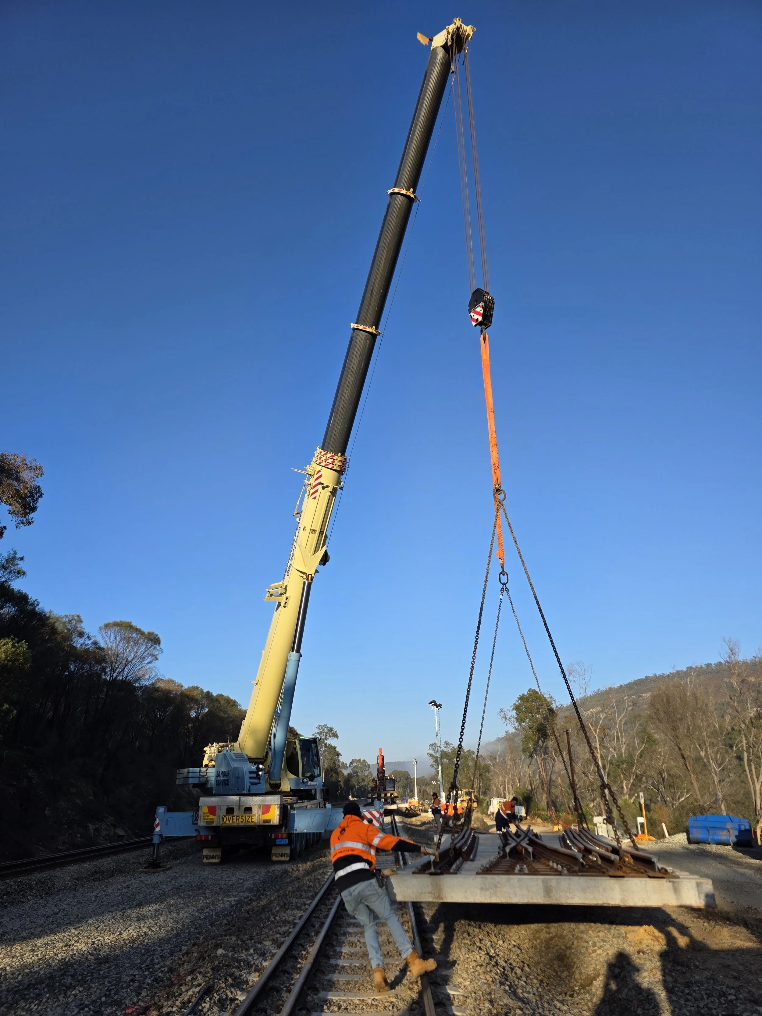 Construction workers guide a large concrete slab onto railroad tracks with a crane lifting it from a flatbed truck, under a clear blue sky.