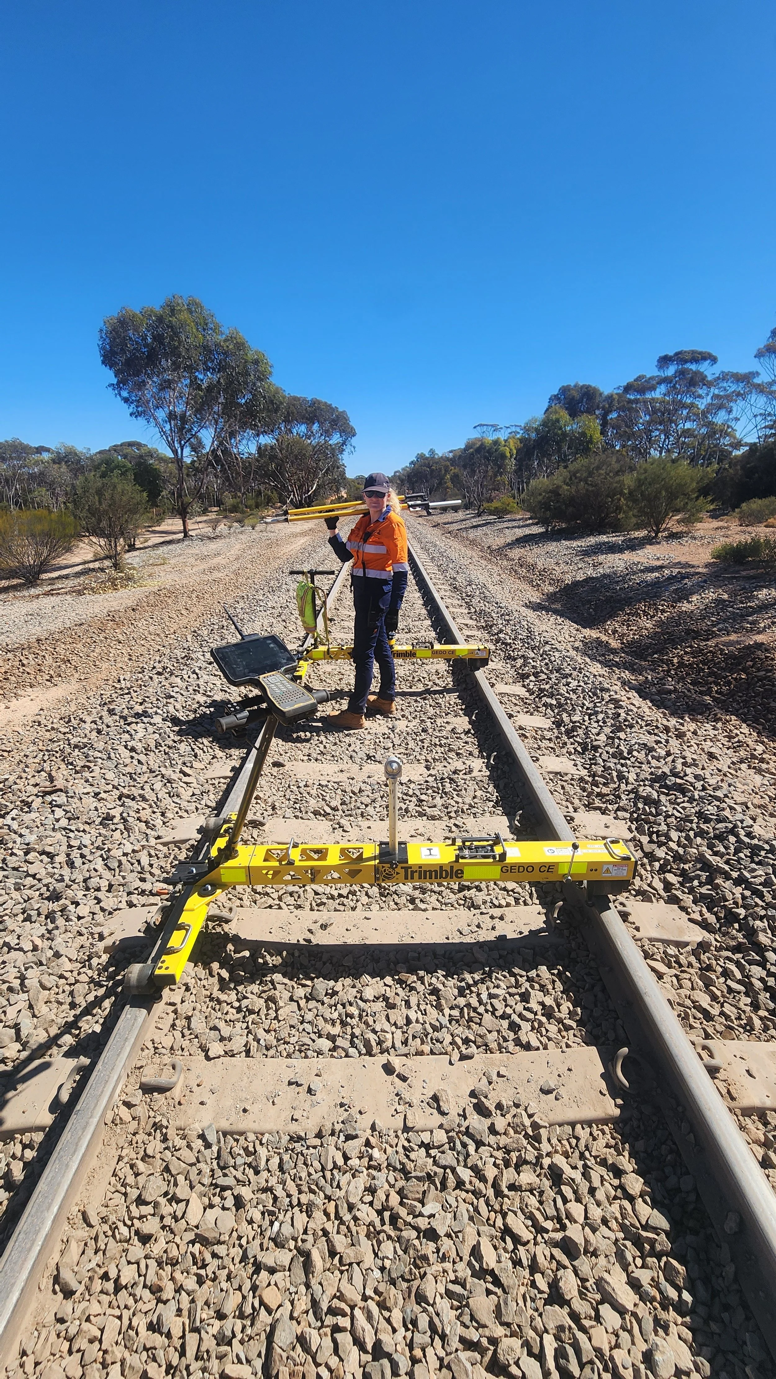 Person in orange jacket and dark pants working on a railroad track in a rural area with trees and a clear blue sky, using surveying equipment.