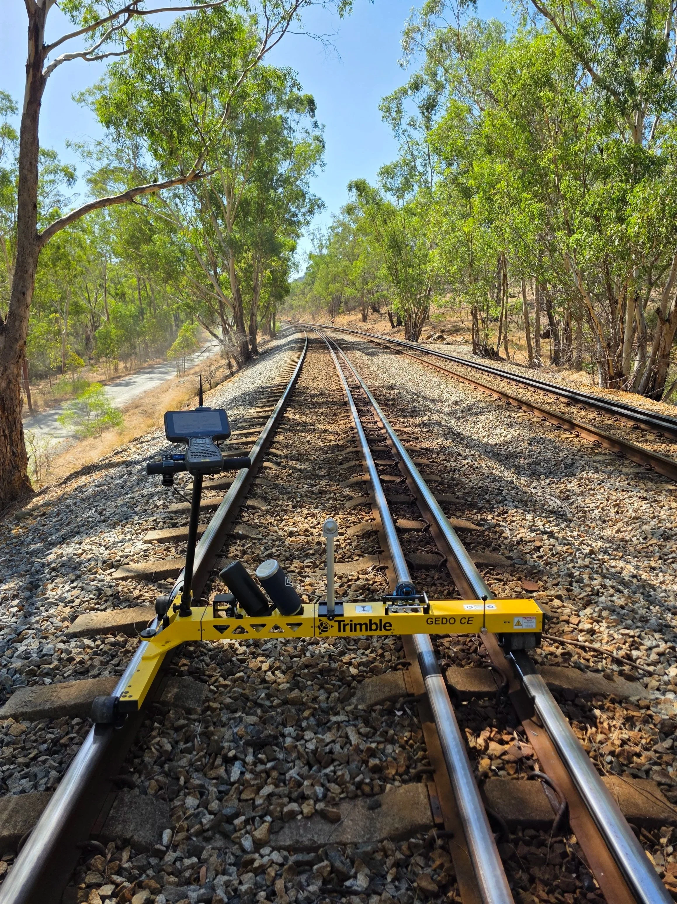 A railway track with trees on both sides, a surveying instrument on the track, and a bright blue sky.