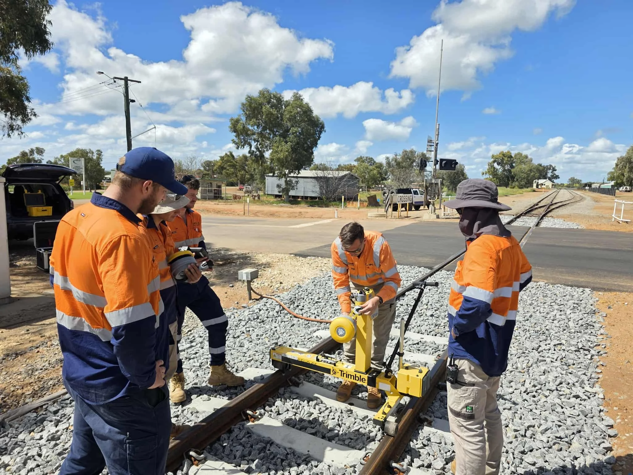 Group of railway workers in orange safety uniforms inspecting and working on train tracks outdoors under a blue sky with clouds.