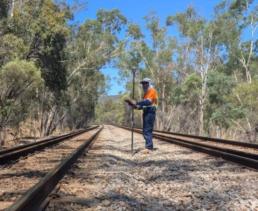 A man in safety gear, including a hat, sunglasses, and a high-visibility orange vest, stands on railroad tracks in a rural, wooded area with blue sky overhead. He is holding a measuring pole and appears to be inspecting the tracks.