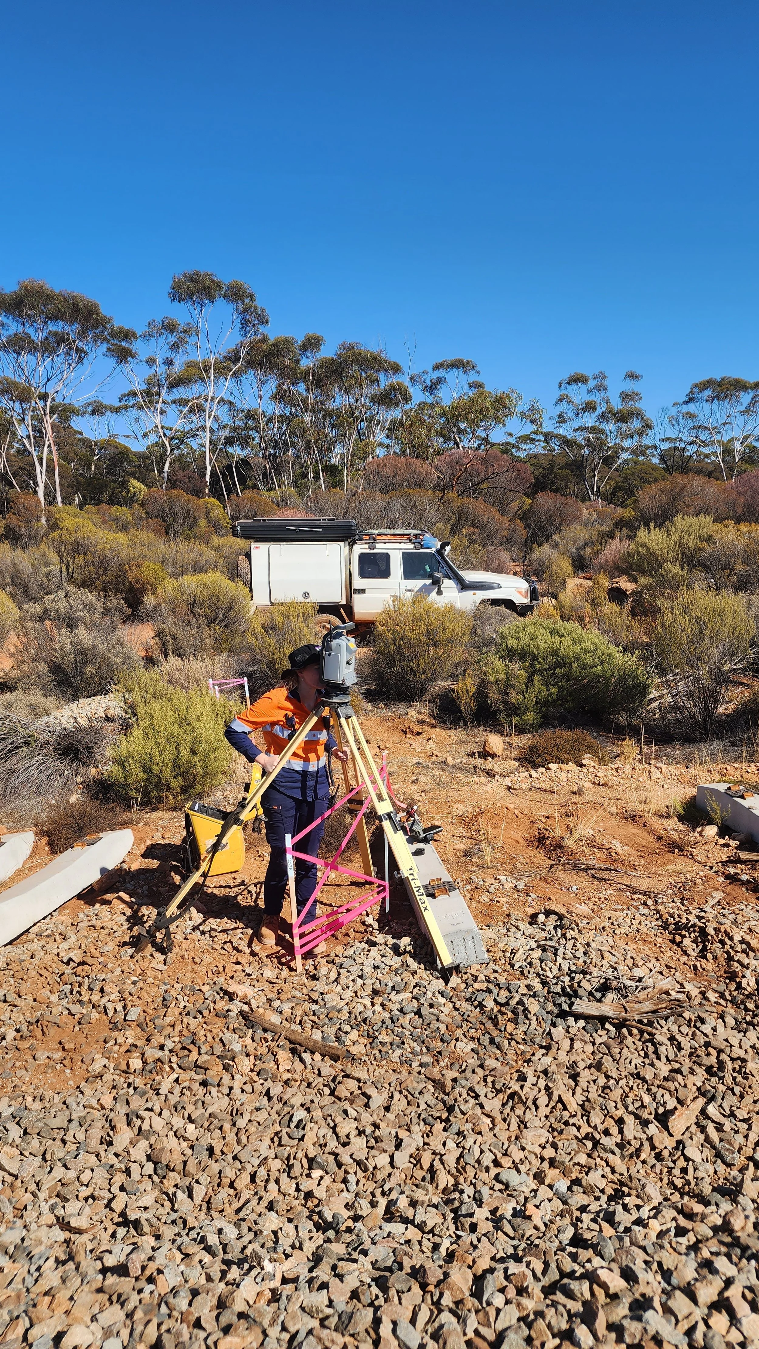 A person working on a surveying project with a theodolite on rocky ground in a desert-like landscape, with bushes and trees, and a white utility vehicle in the background under a clear blue sky.