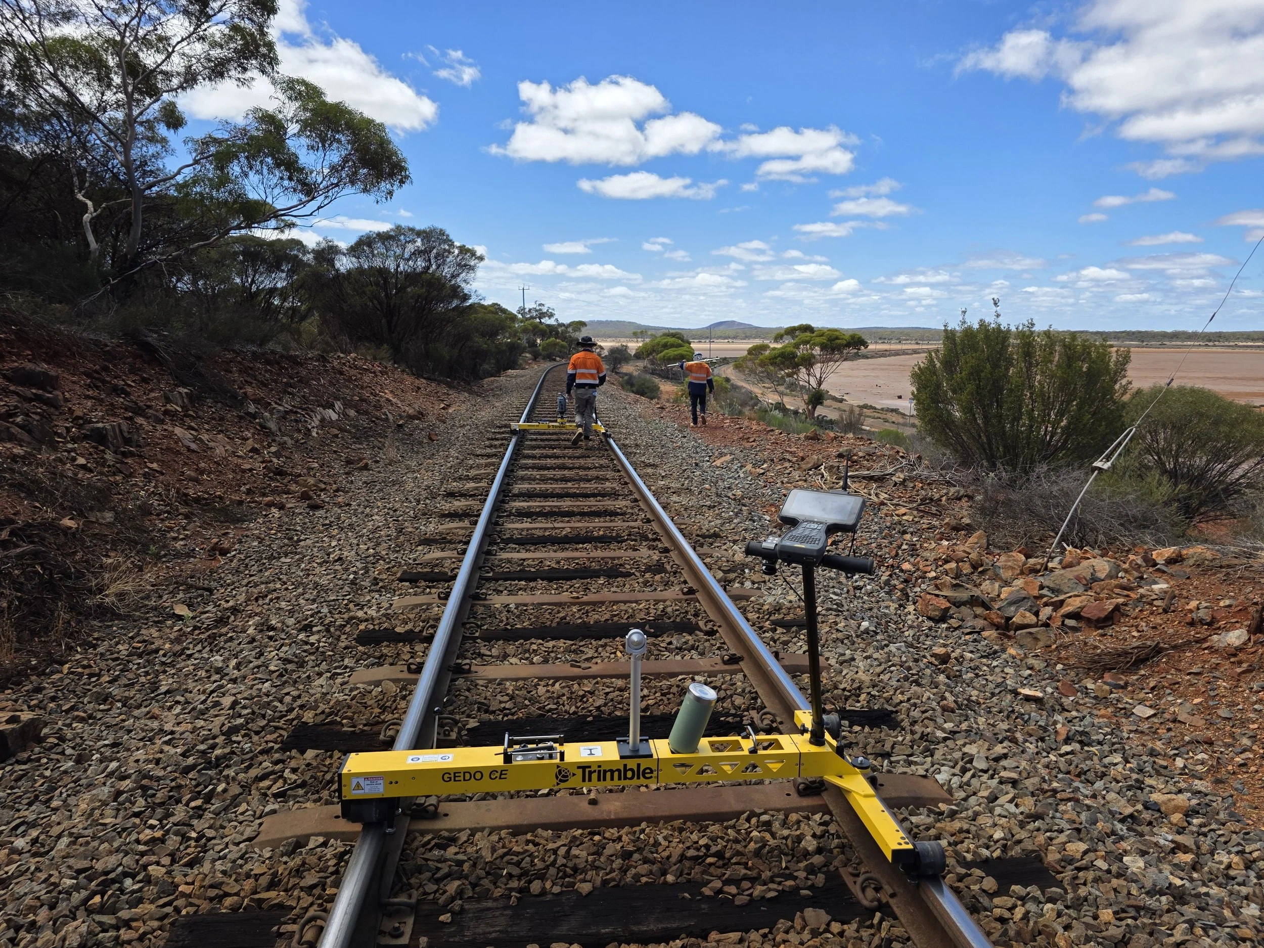 Workers in orange safety vests installing or repairing railway tracks on a rural landscape with scattered trees and a partly cloudy sky.