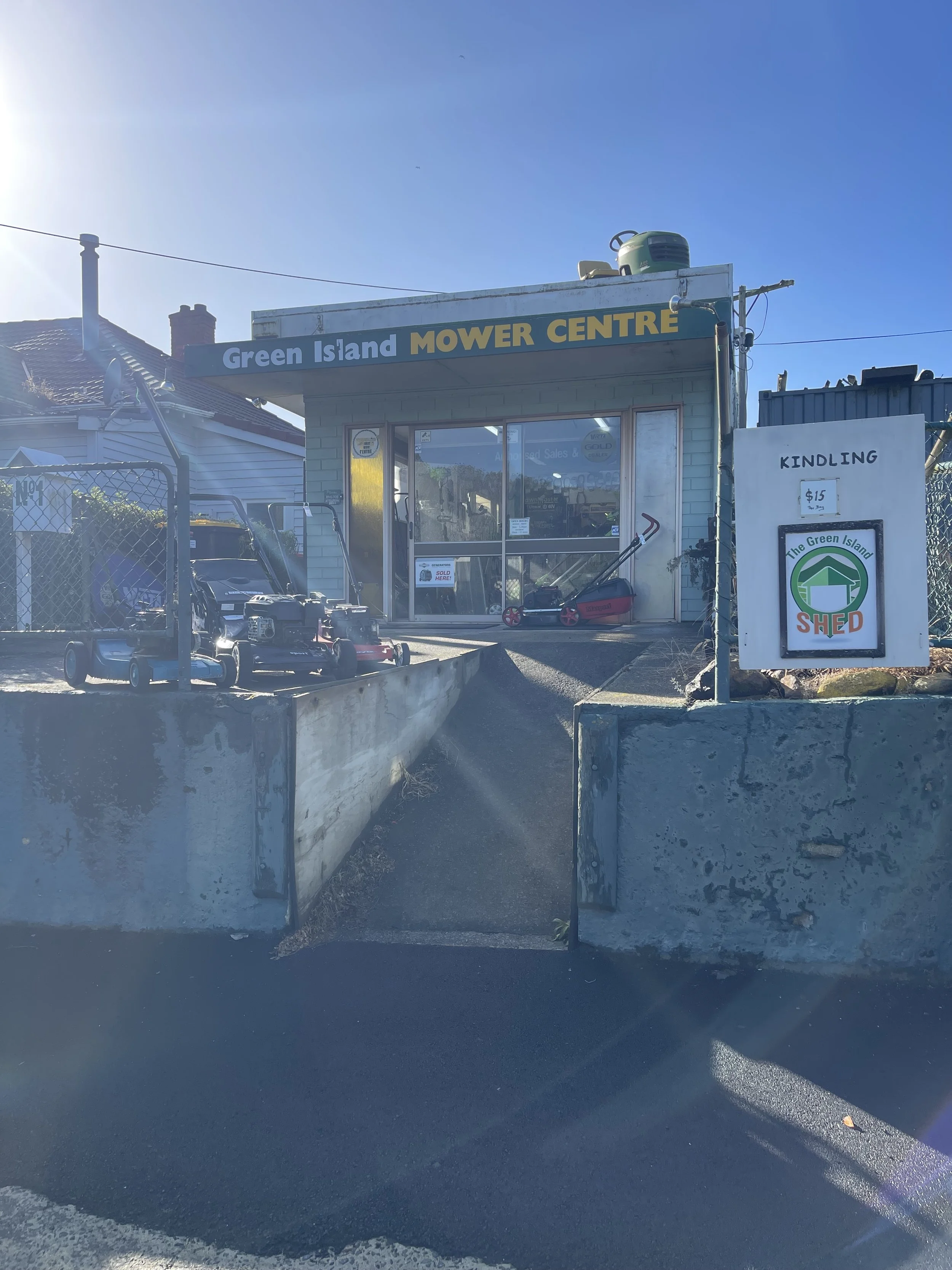 The Green Island Mower Centre storefront with lawn mowers outside, a sign offering kindling for $15, and a small shed sign. The building is on a raised platform with a ramp, and the background includes neighboring houses and a clear blue sky.