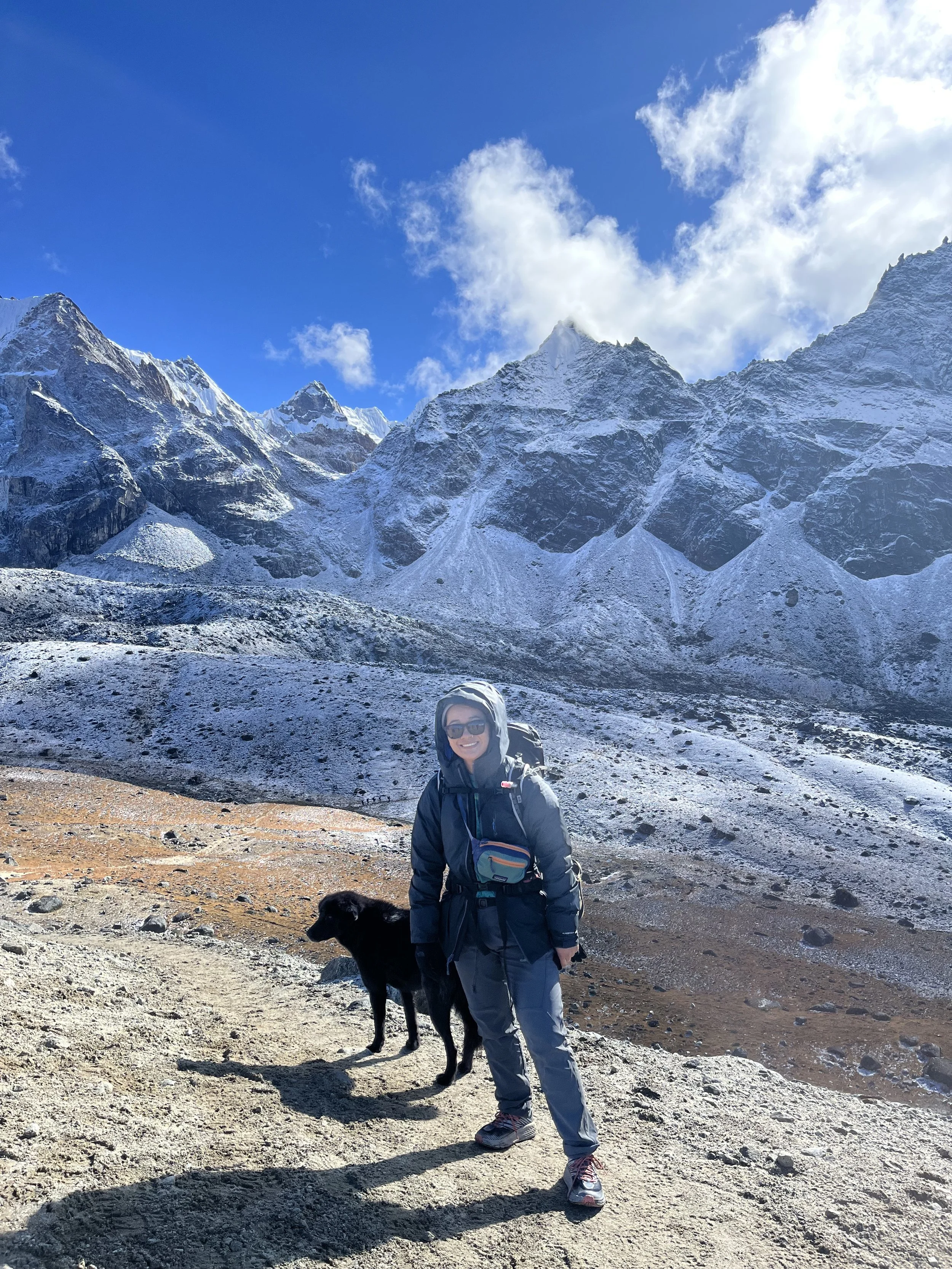 A woman in hiking gear standing with a black dog in a mountainous landscape with snow-capped peaks and a clear blue sky. Nurse Practitioner who loves hiking and mountains.