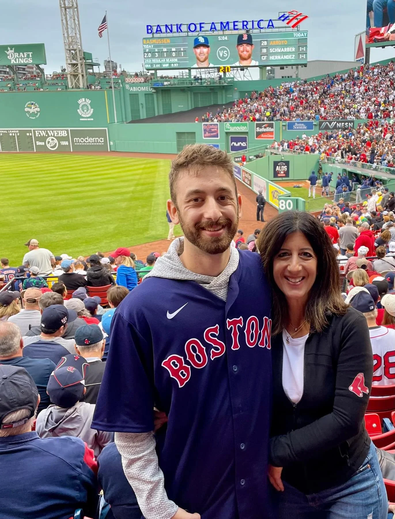 A man and woman smiling, wearing Boston Red Sox apparel, at Fenway Park during a baseball game.