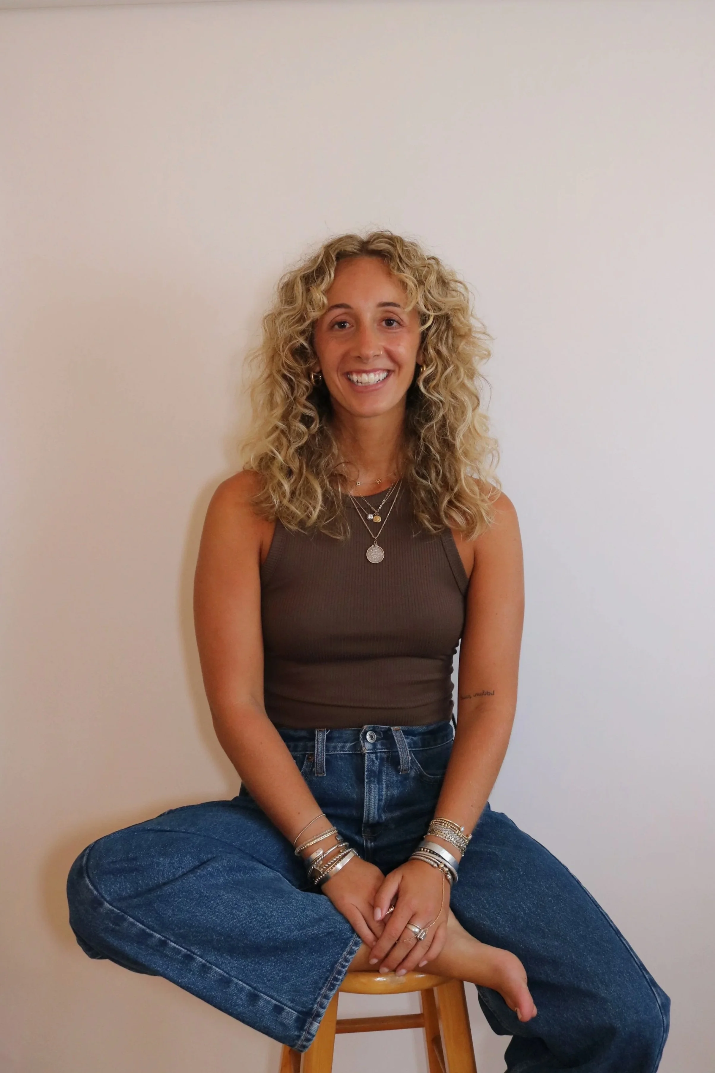 A young woman with curly blonde hair, smiling, wearing a brown sleeveless top, blue jeans, multiple bracelets, necklaces, and rings, sitting cross-legged on a wooden stool against a plain white wall.