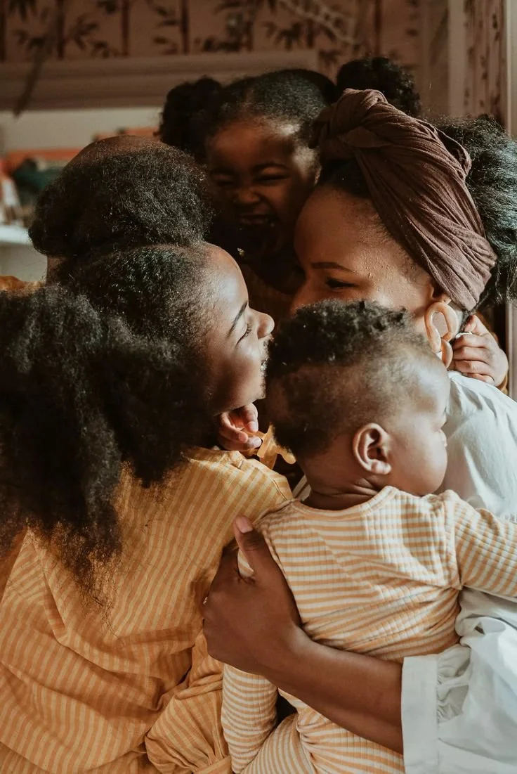 A woman surrounded by four children, all smiling and hugging each other warmly in a cozy indoor setting.