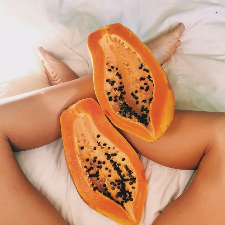 A person sitting on a bed with a sliced papaya fruit on their lap and legs, showcasing orange flesh and black seeds.