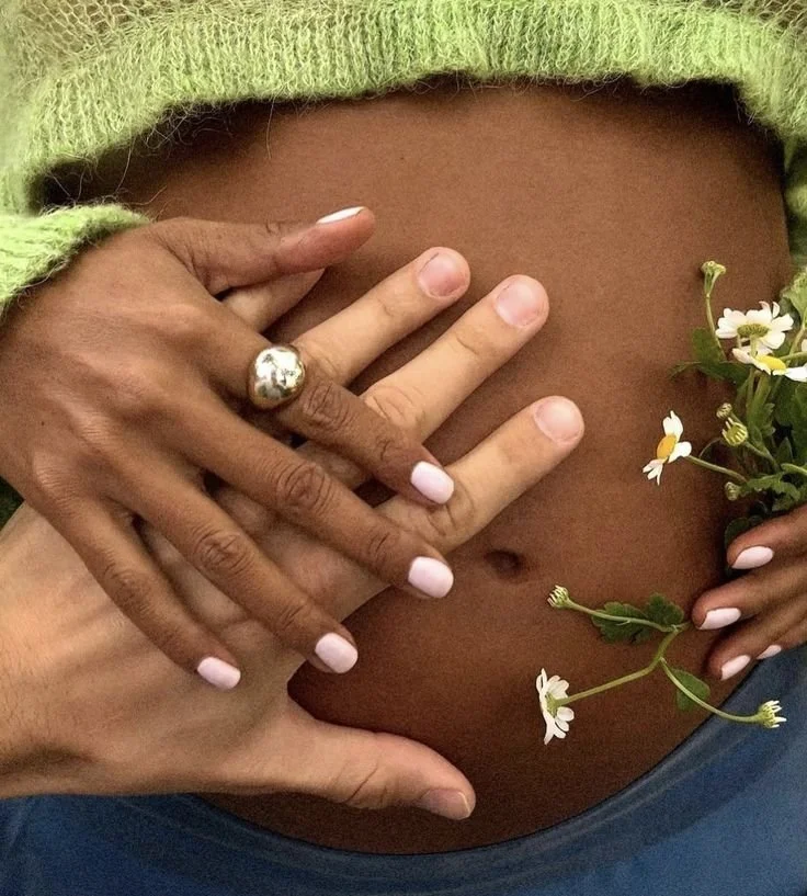 Two people gently holding a pregnant woman's belly, with one hand on top, and a small bouquet of flowers in her hand.