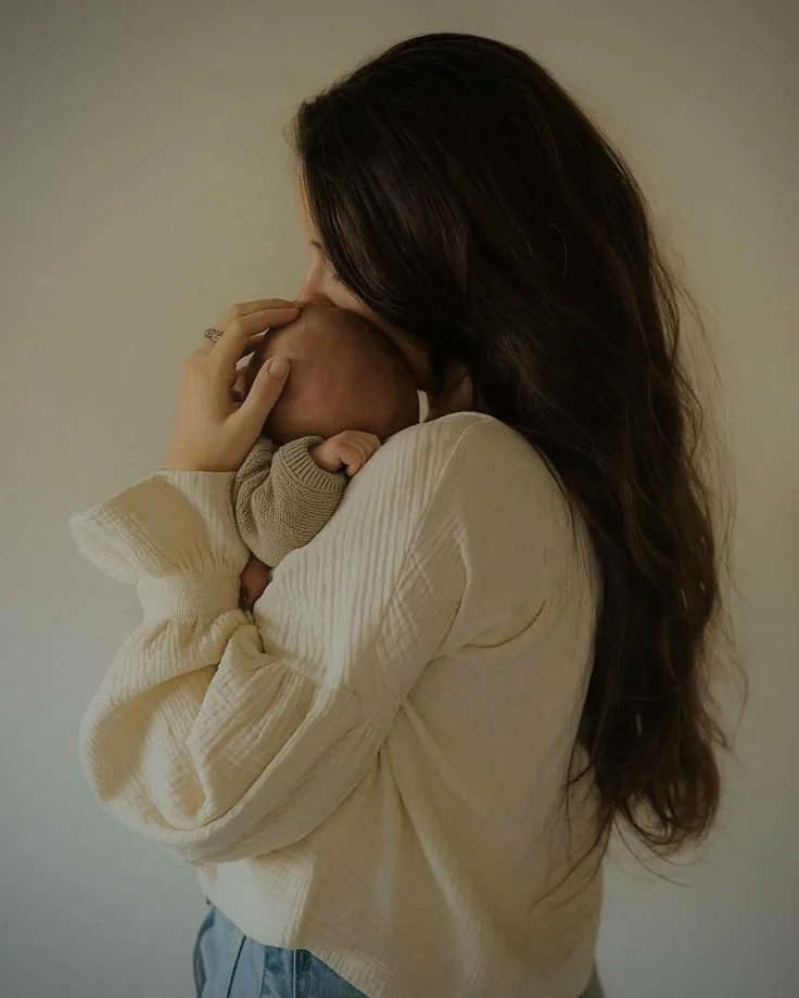 A woman holding a baby close to her face indoors with a neutral background.