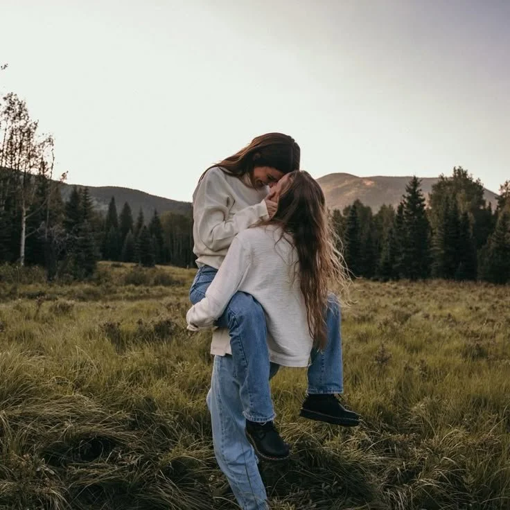 Two women sharing an intimate moment outdoors in a grassy field with trees and mountains in the background.