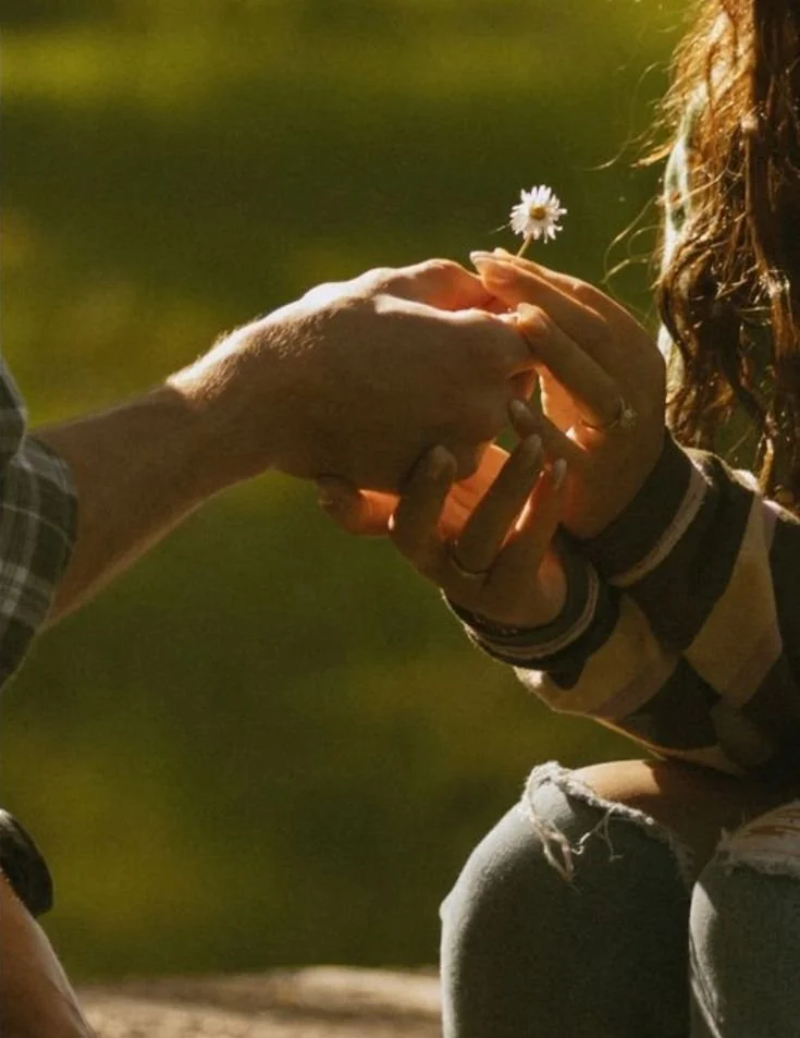 A person is handing a coin to another person, who is holding a small flower, in an outdoor setting with green background.