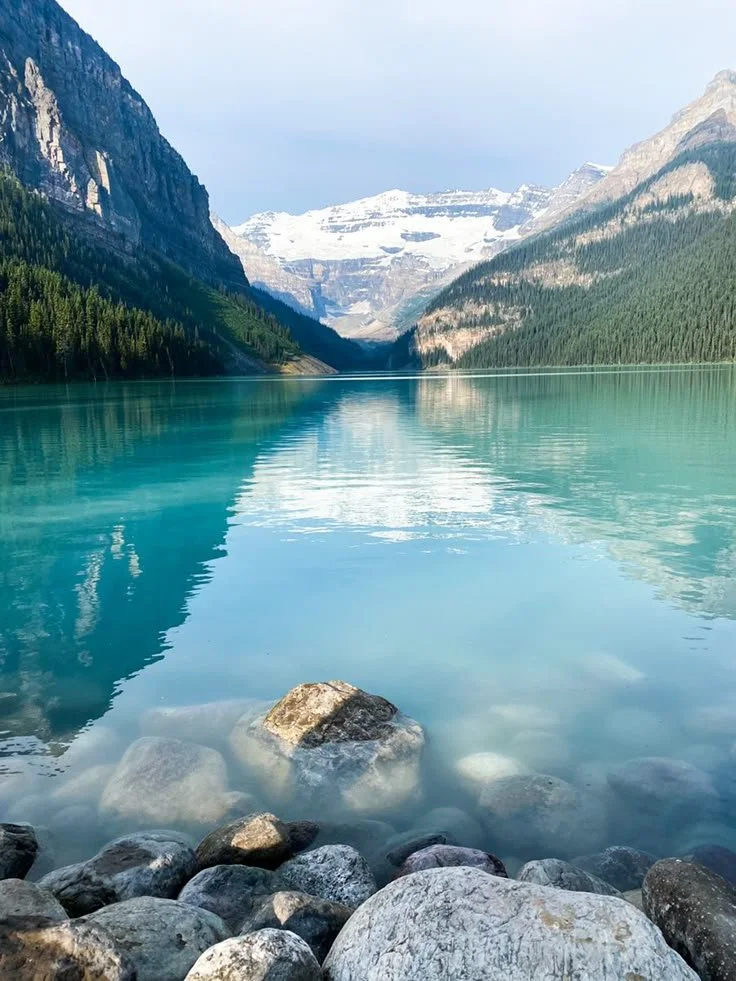 A tranquil mountain lake with clear blue water, surrounded by forested mountains and snow-capped peaks in the distance. Large rocks line the shoreline in the foreground.