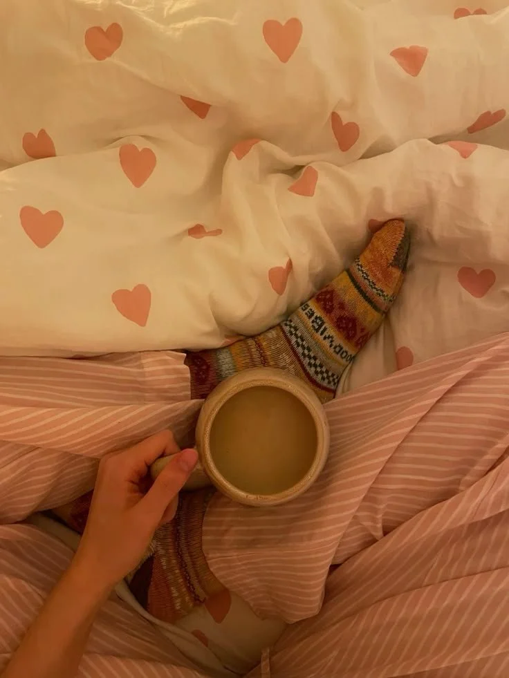 Person sitting on a bed with pink and white striped sheets and a heart-printed comforter, holding a beige mug with a yellowish drink, wearing colorful patterned socks.