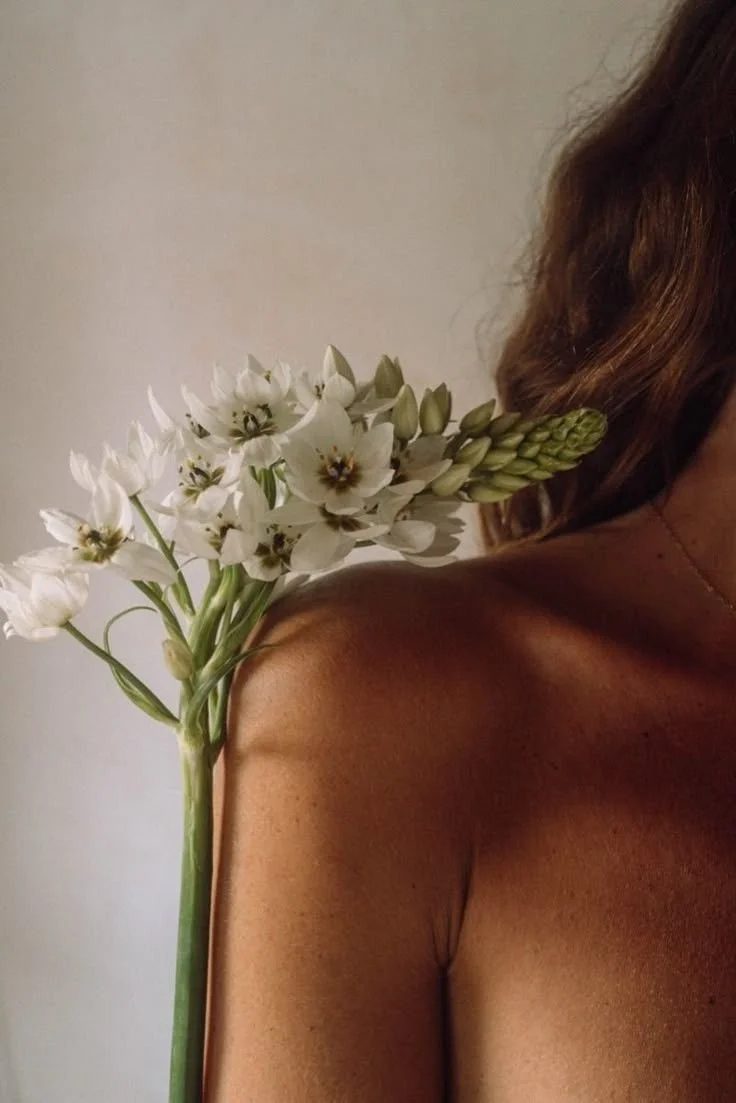 Close-up of a woman's shoulder with a bouquet of white flowers resting on it, against a plain background.