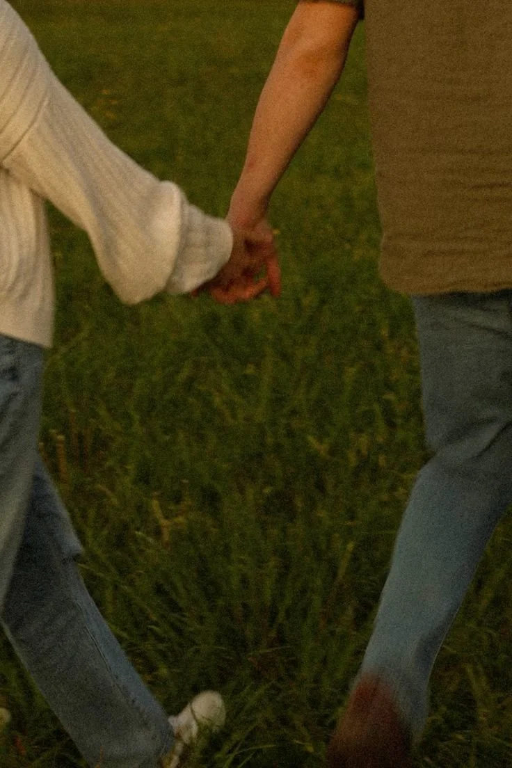 Two people holding hands, walking through a grassy field at night.