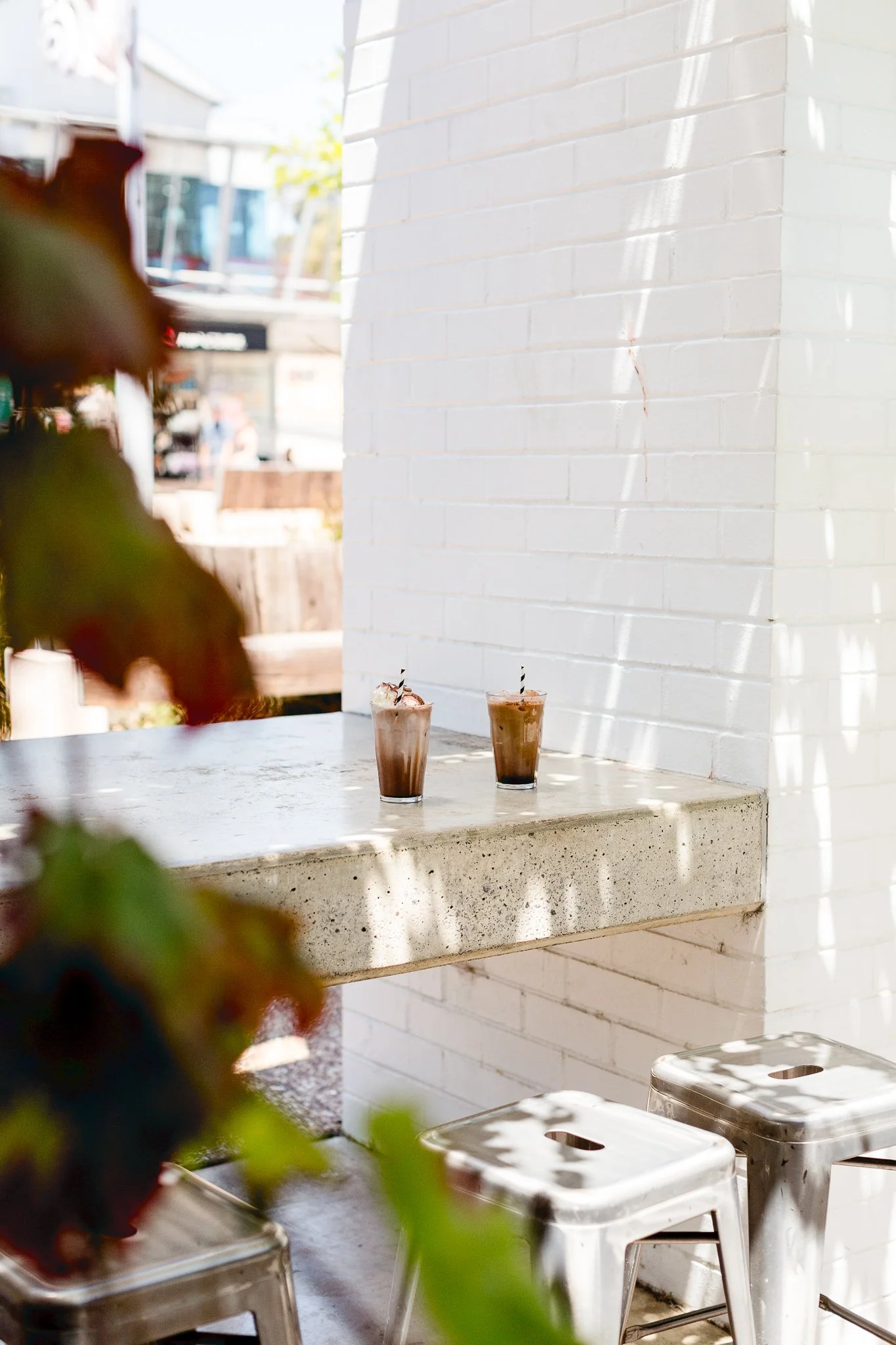 Two iced coffee drinks with straws placed on a concrete bar table outside a cafe, against a white brick wall with sunlight and shadows.