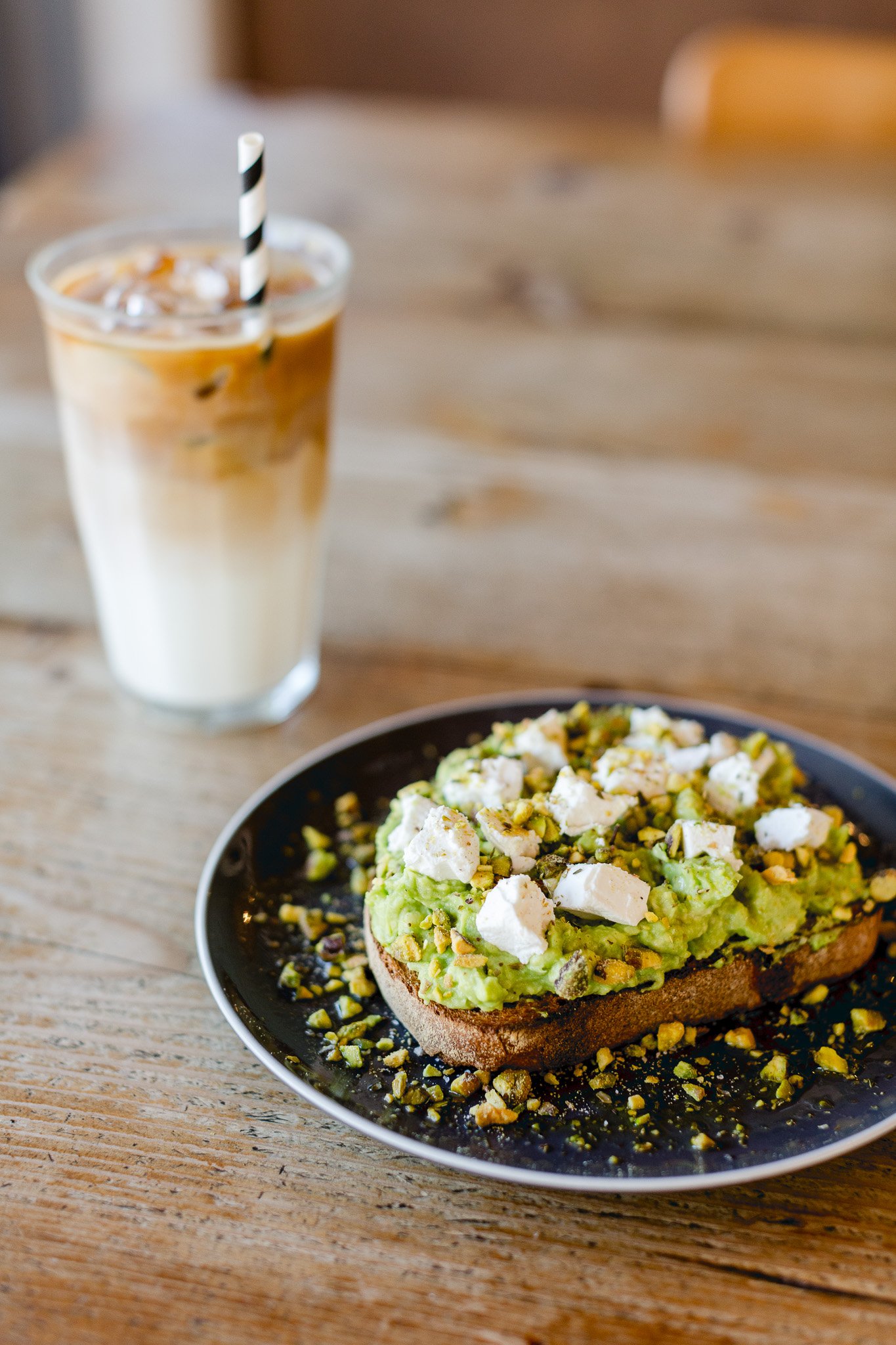 Toasted bread topped with avocado, crumbled cheese, and chopped pistachios on a black plate, with a glass of iced coffee in the background on a wooden table.