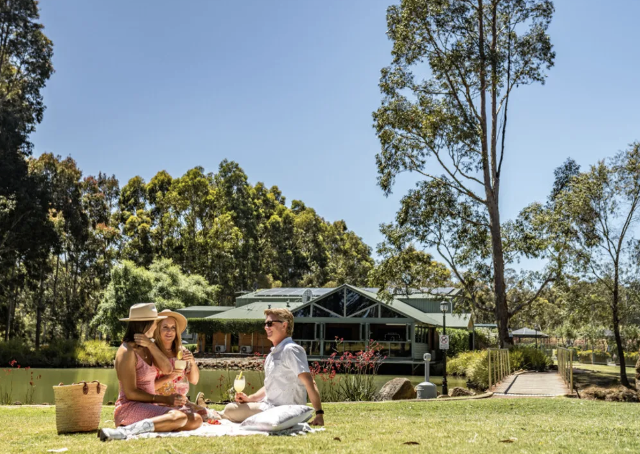 Three friends having a picnic near a pond with a building in the background on a sunny day.