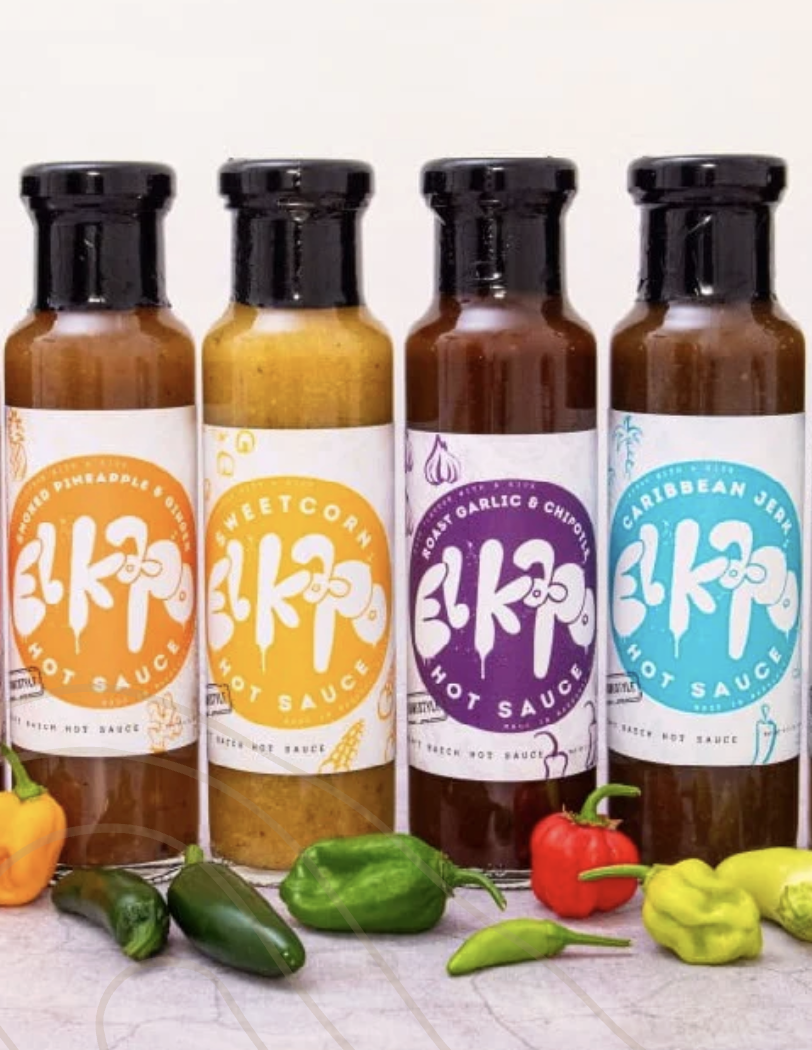 Four bottles of hot sauce with colorful labels standing in a row, surrounded by small peppers and vegetables on a white surface.