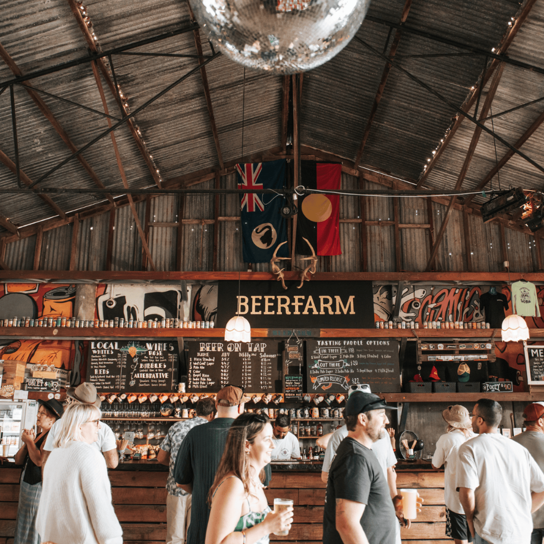 People gather inside a rustic beer farm with a corrugated metal roof, wooden beams, and colorful mural walls, enjoying drinks and socializing at the bar.