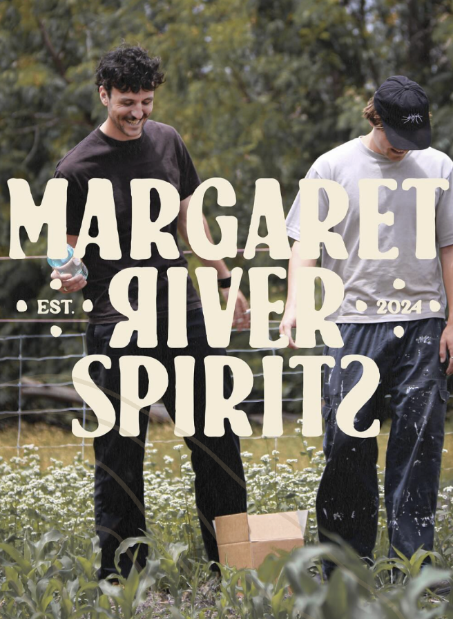 Two young men are standing outdoors in a field of plants, smiling and looking down. One is holding a water bottle, and both are dressed casually. Large text overlay reads "MARGARET RIVER SPIRITS" with "EST. 2024" on the sides. The scene appears to be cheerful and relaxed.