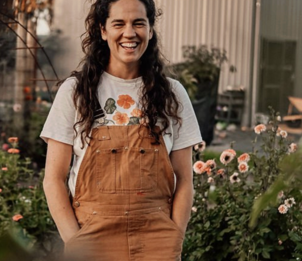 A woman with curly hair smiling outdoors among pink flowers, wearing a white t-shirt with floral designs and orange overalls.