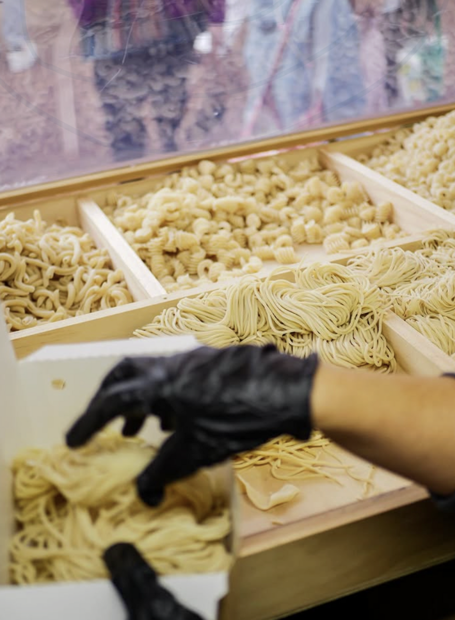 A person wearing black gloves handling fresh pasta in a display case at a grocery or pasta shop.