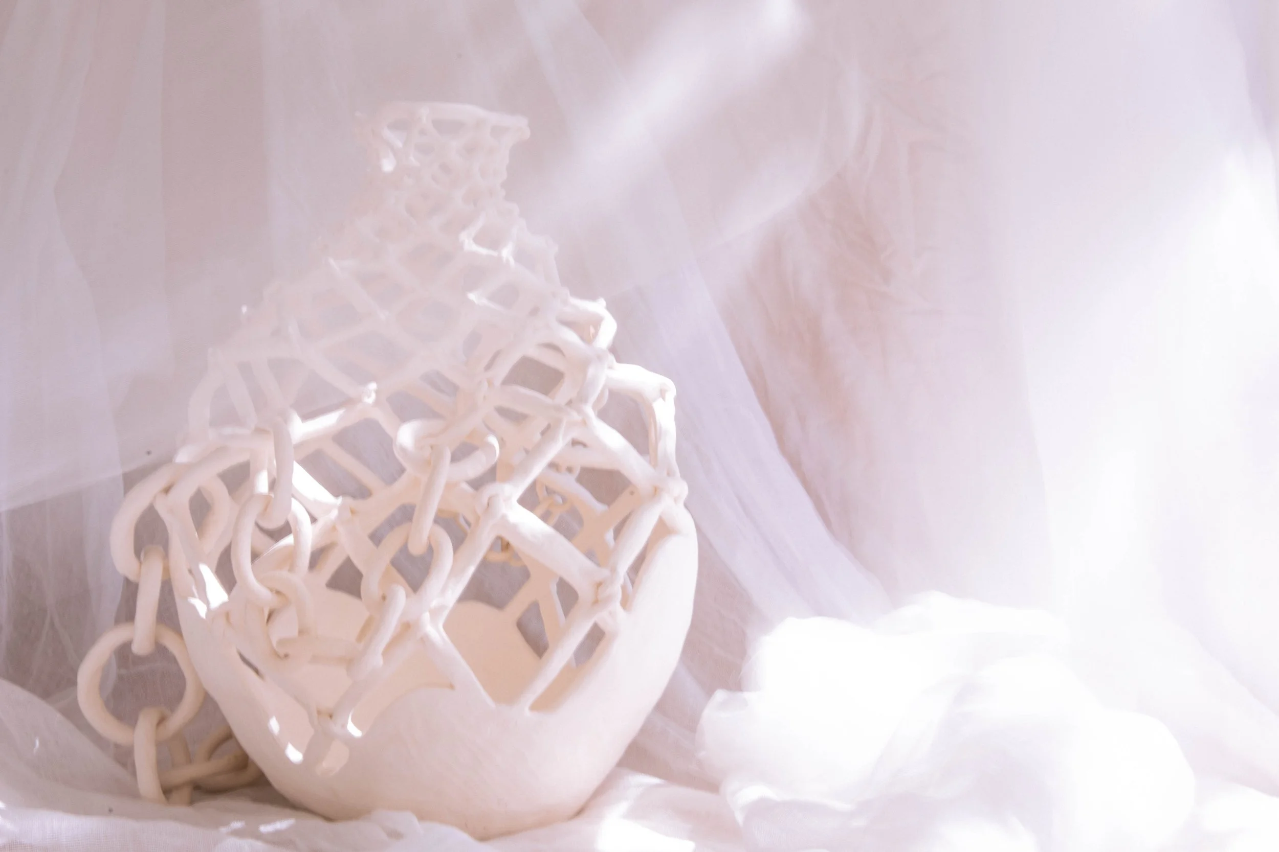 White ceramic vase with an intricate lattice design, set against a soft, light background with translucent drapery and fabric.