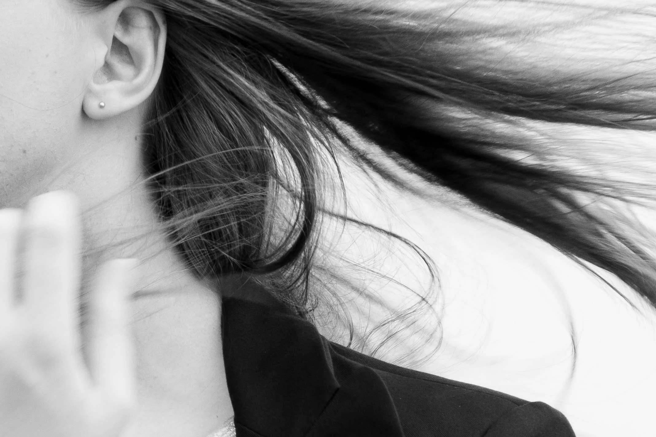 Close-up of a woman's face and hair, with a focus on her ear, wearing a small pearl earring, and her hand near her face. She has long hair flowing to the right.