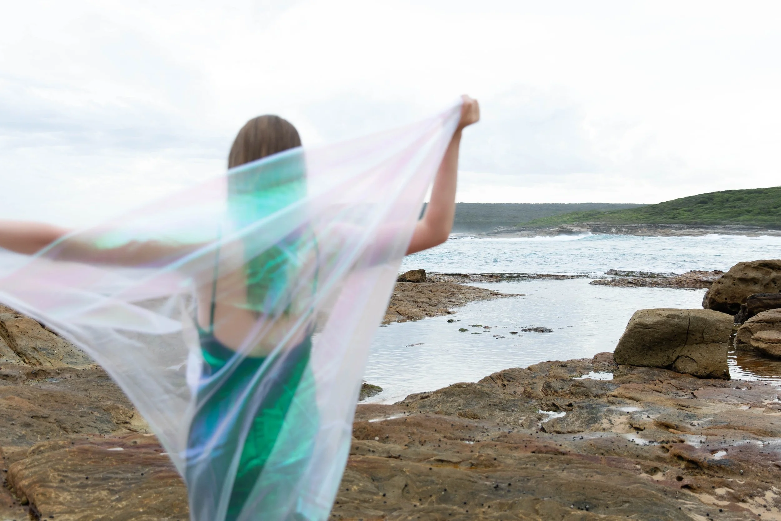 Woman standing on rocky shoreline holding a sheer, iridescent veil with arms outstretched, overlooking the ocean with waves and a distant green hillside under a cloudy sky.