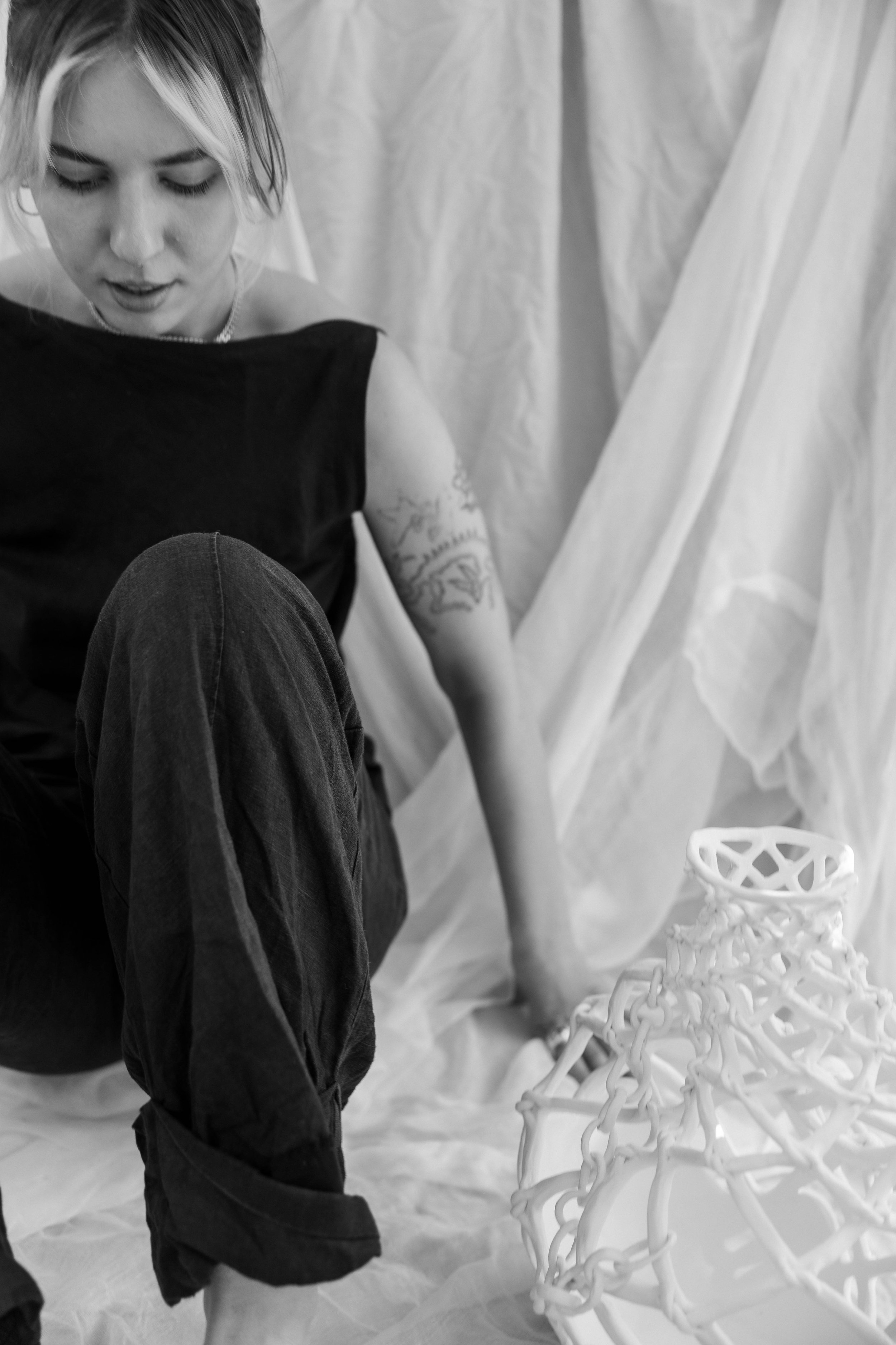 A woman with short blonde hair, tattoos on her arm, wearing a sleeveless black top, is sitting on the floor with a draped fabric background. There is a white, intricate, lattice-like ceramic sculpture in front of her.