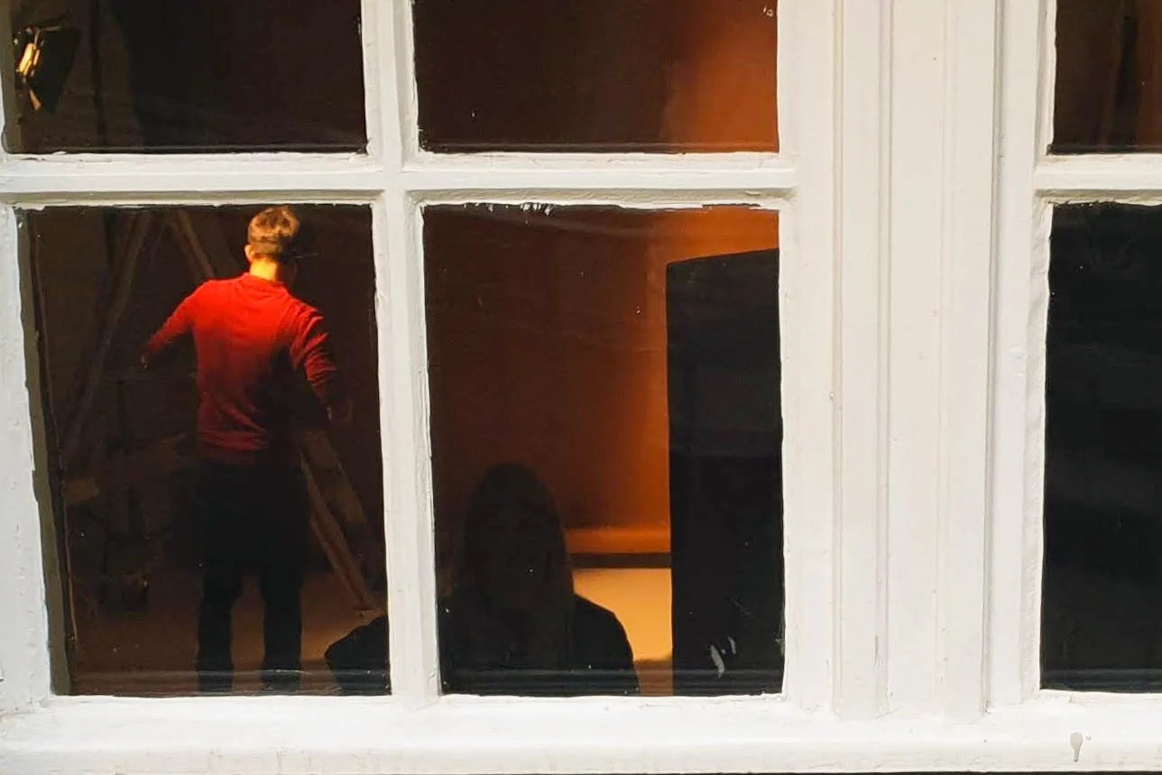 A person wearing a red jacket, taking a photo inside a photography studio with orange lighting, seen through a window.