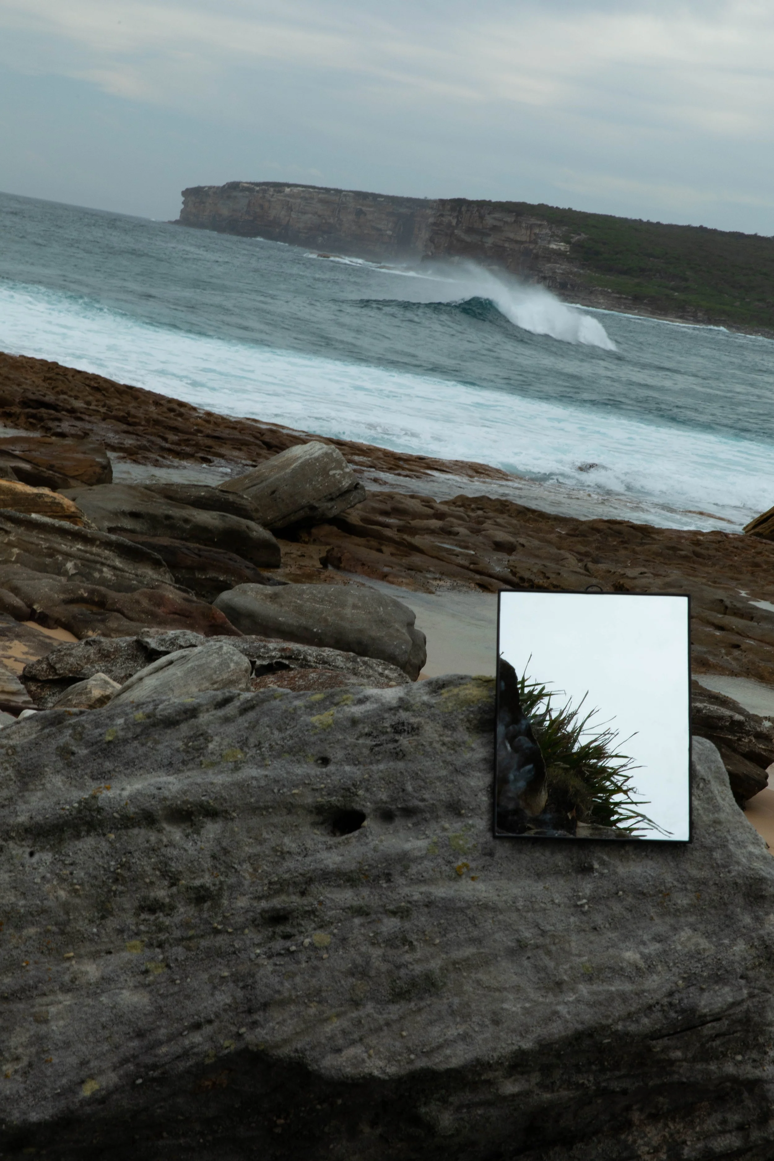 Rocky shoreline with ocean waves, a distant cliff, and a small mirror reflecting coastal plants.