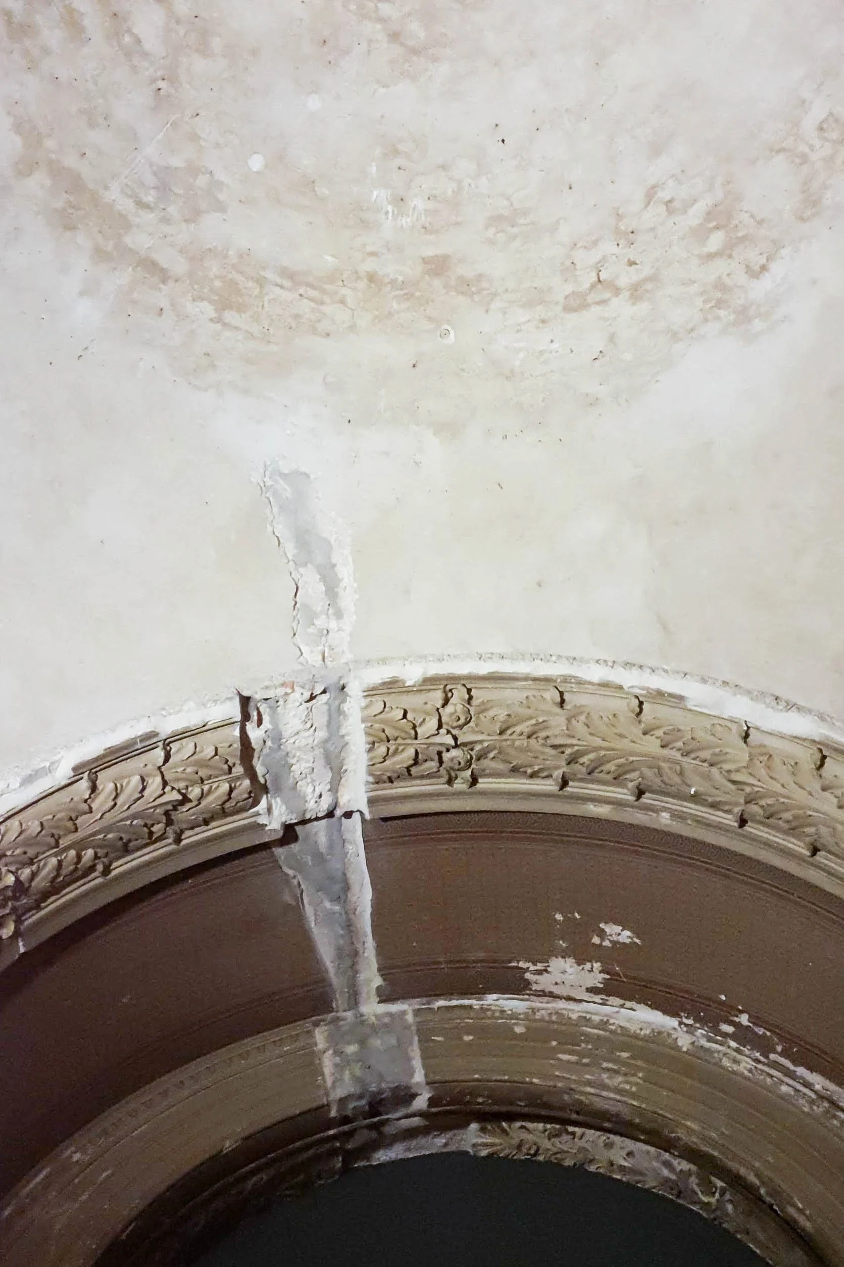 Close-up view of a cracked and damaged decorative ceiling molding and plaster with a visible vertical crack in the ceiling.