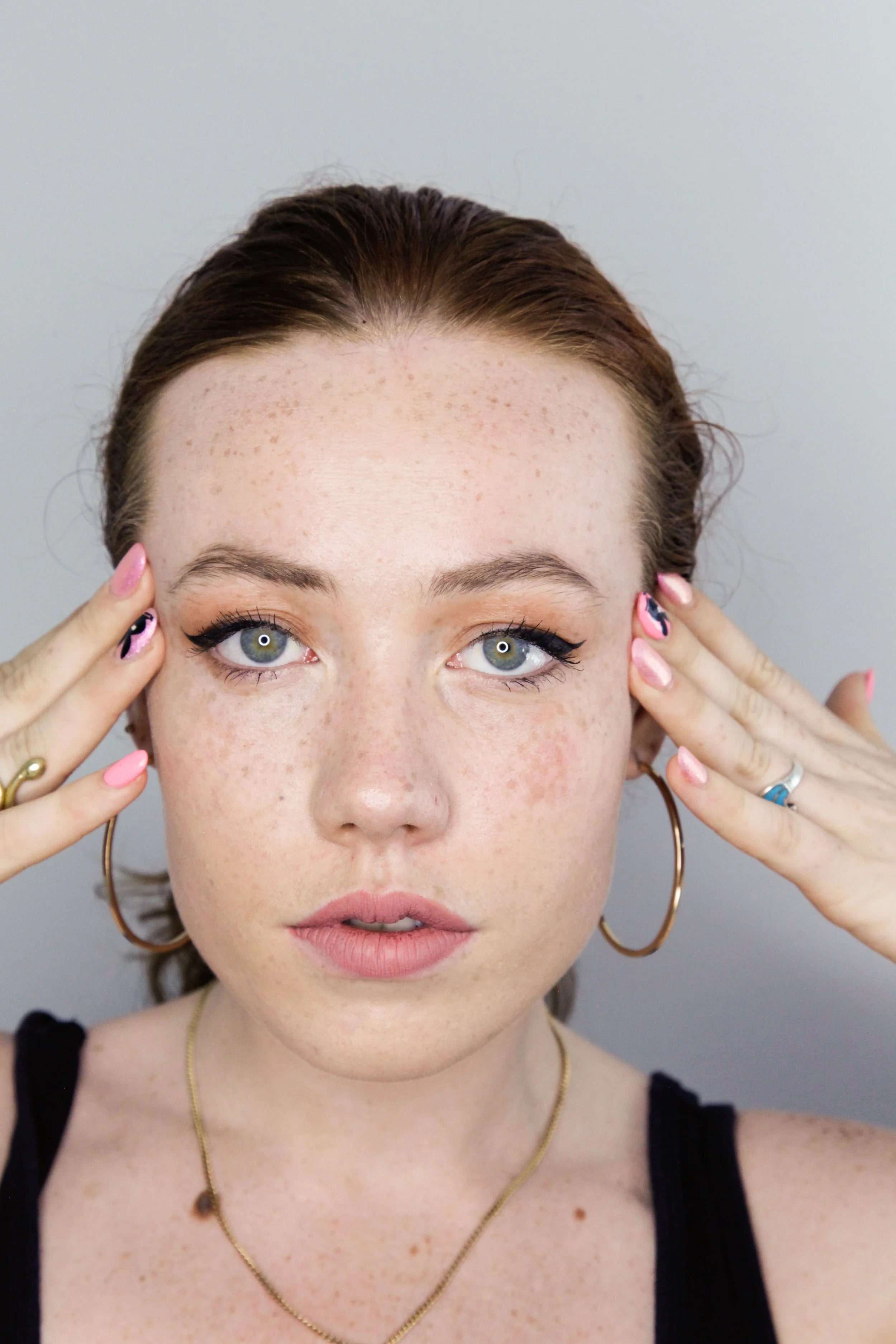 Close-up portrait of a young woman with blue eyes, freckles, and red hair, touching her temples with her fingers. She has makeup, earrings, and a necklace, wearing a black tank top against a plain grey background.