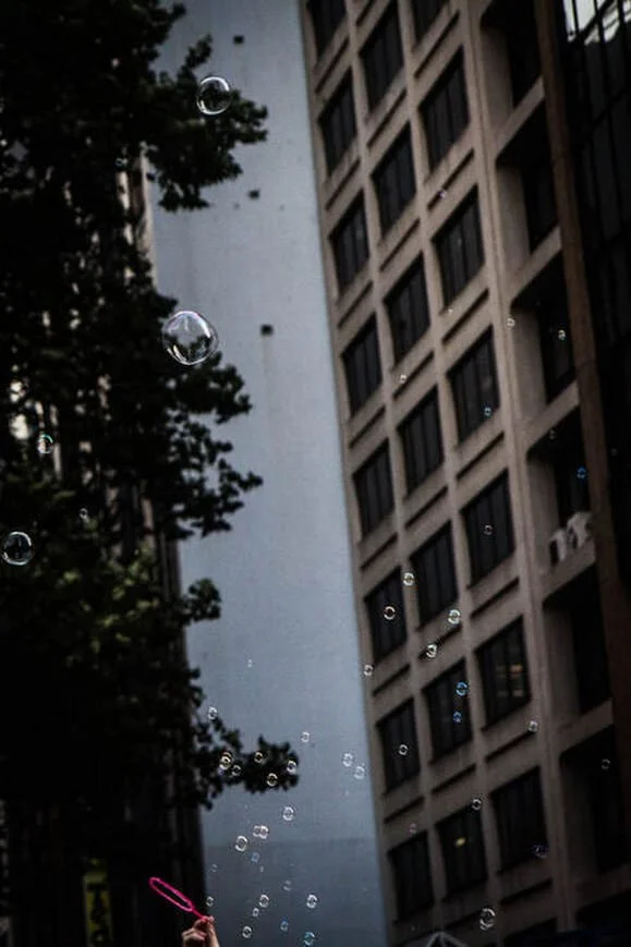 A person in the lower left corner holding a pink bubble wand, blowing bubbles into the air. Tall buildings and a tree are in the background on an urban city street.