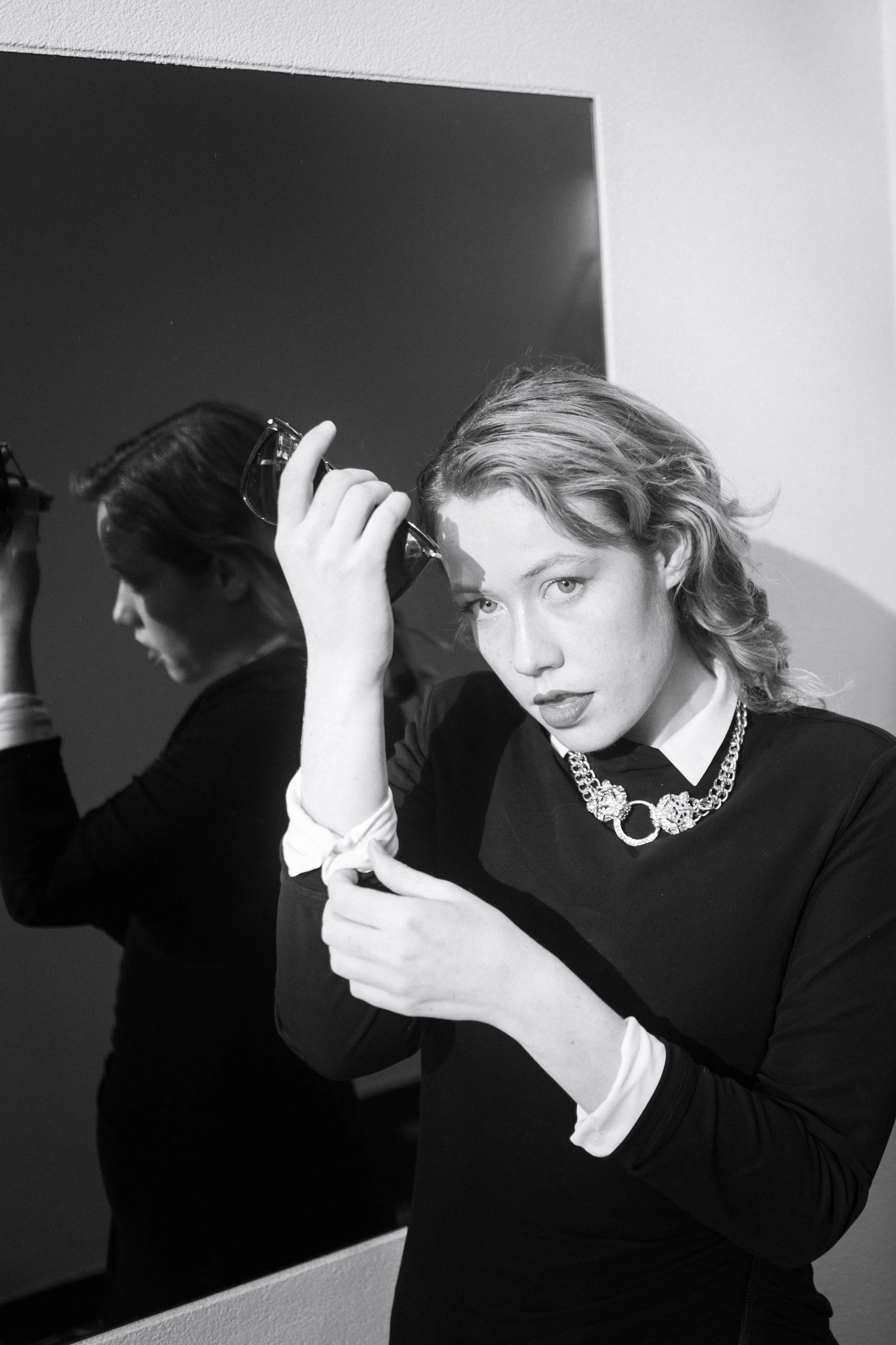 A woman with wavy hair holding her hands above her head, looking at the camera, with her reflection visible in a mirror behind her.