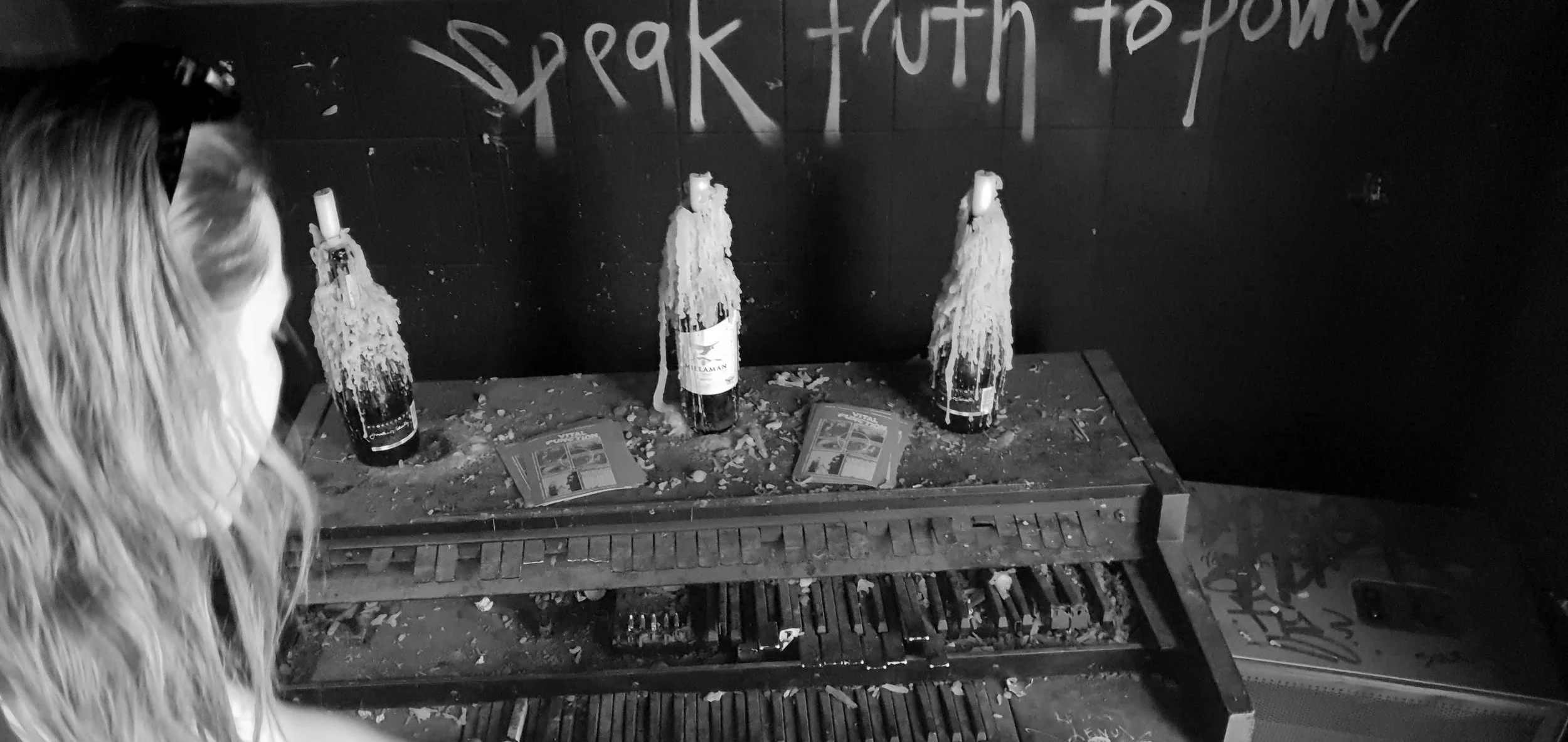 A vandalized piano with three bottles containing melted candles, and scattered debris, with graffiti on the wall behind that reads 'Speak truth to power'.