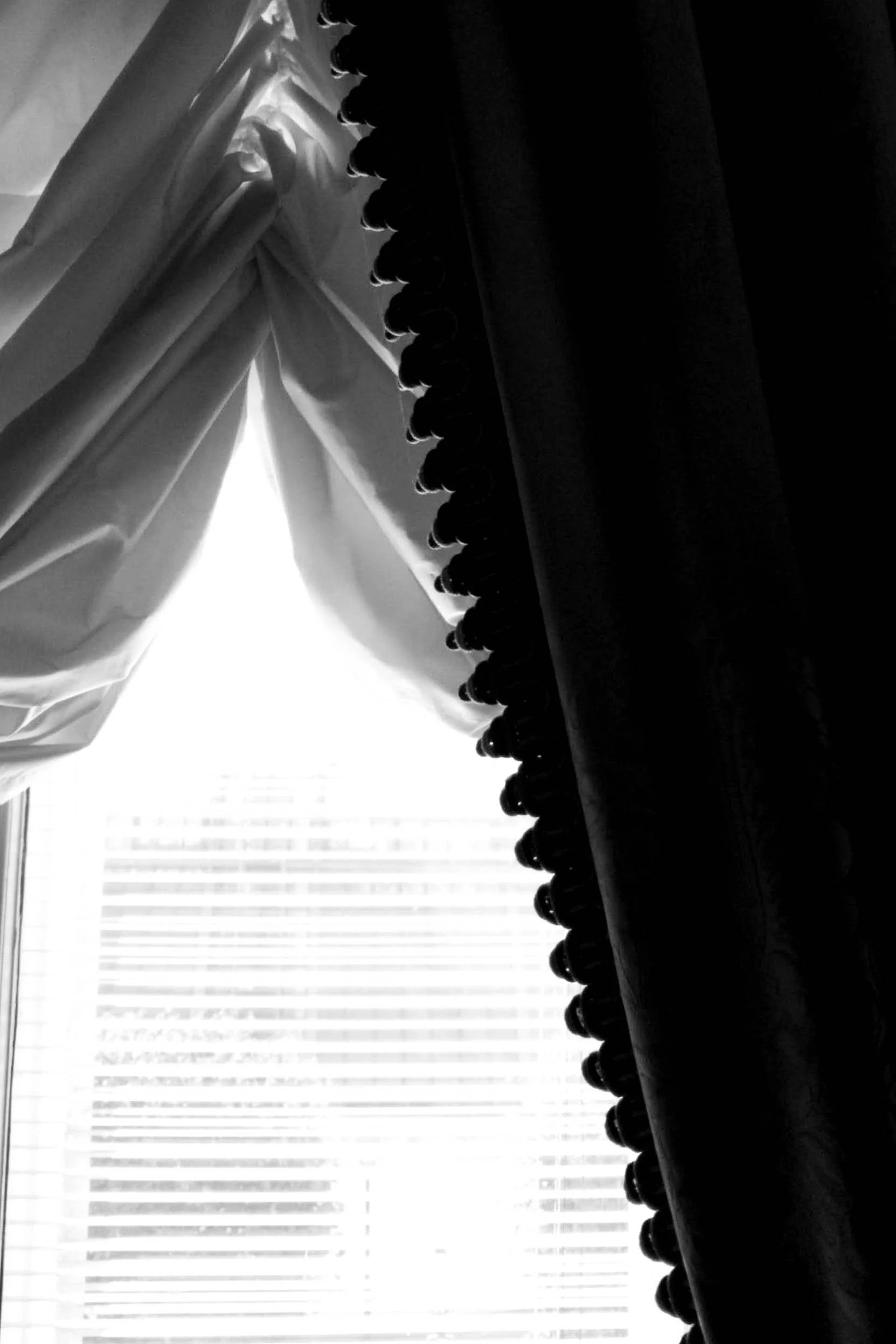 Black and white photo looking through window with curtains and valance, blinds in background.