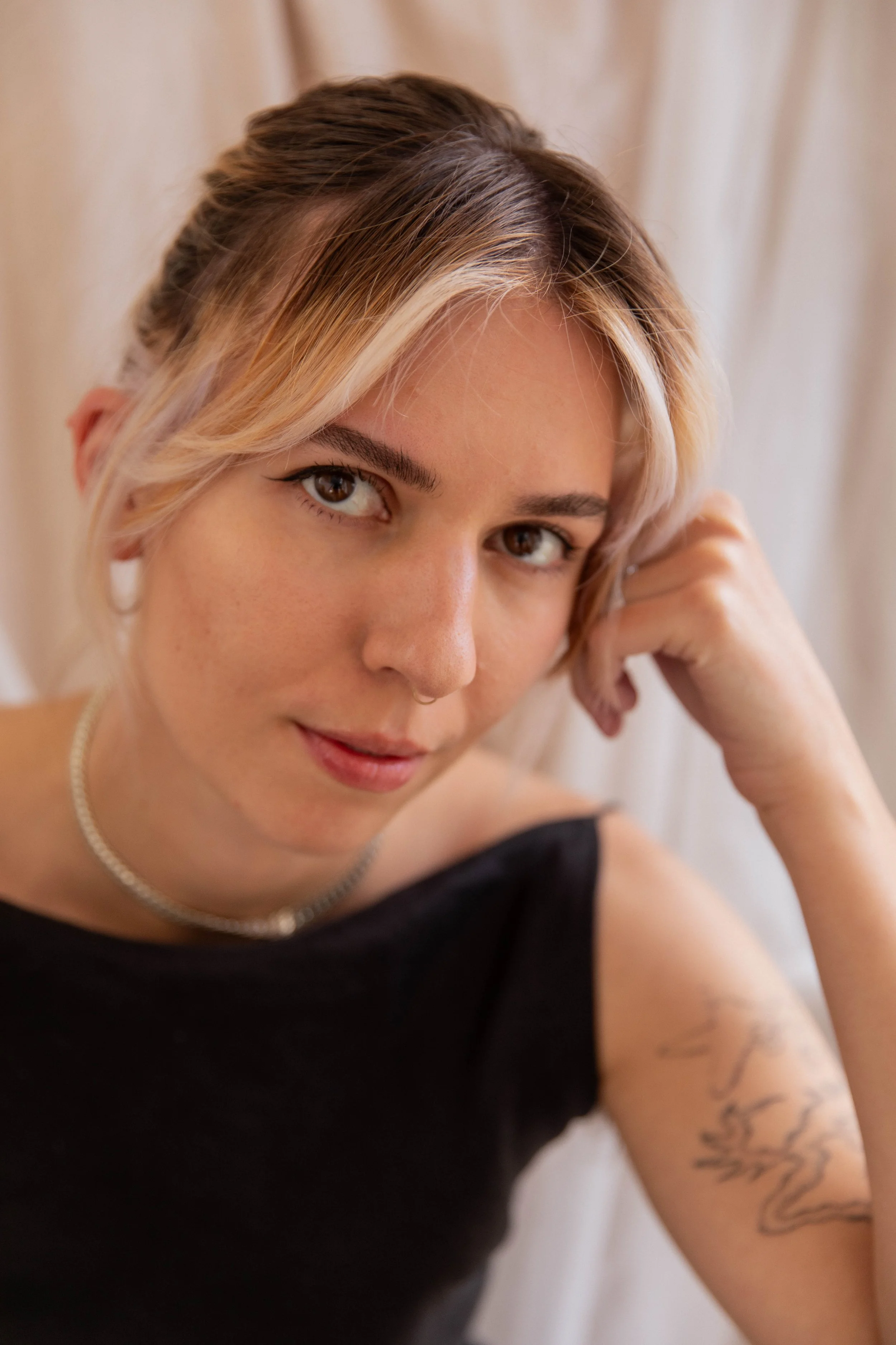 A woman with blonde hair styled in a loose updo, wearing a black sleeveless top, a pearl necklace, and a tattoo on her upper arm, sitting in front of a light-colored curtain.