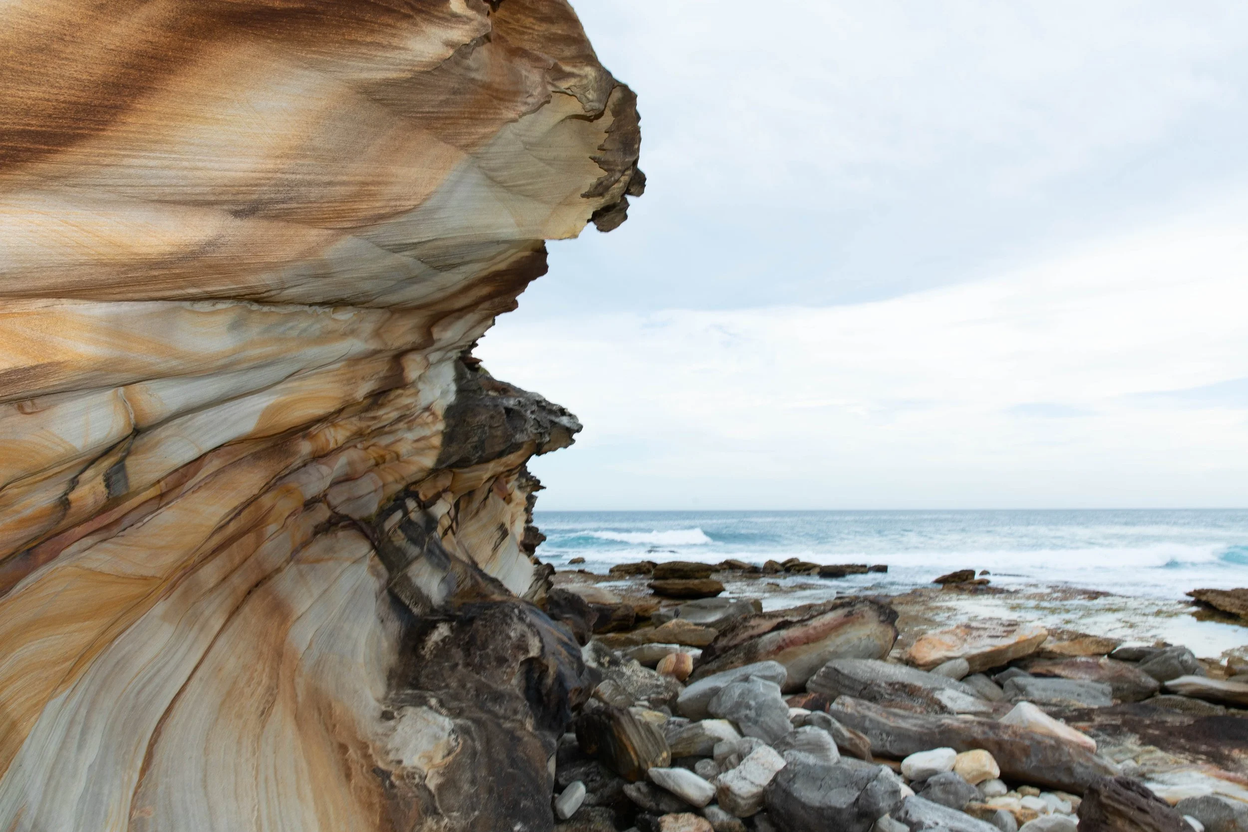 Photo of a rocky coastline with large layered sandstone cliffs on the left and the ocean in the background, under a cloudy sky.