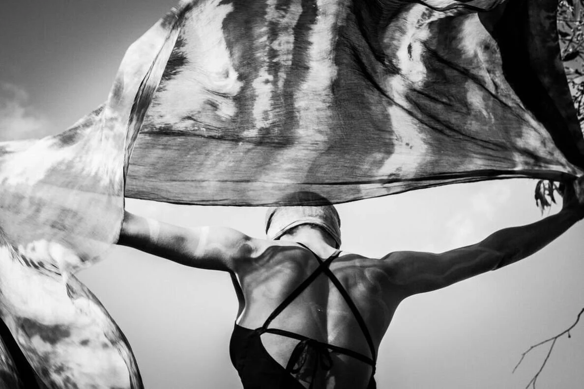 Black and white photo of a woman with arms stretched out, holding a large flowing cloth above her back, with sky and tree branches visible.
